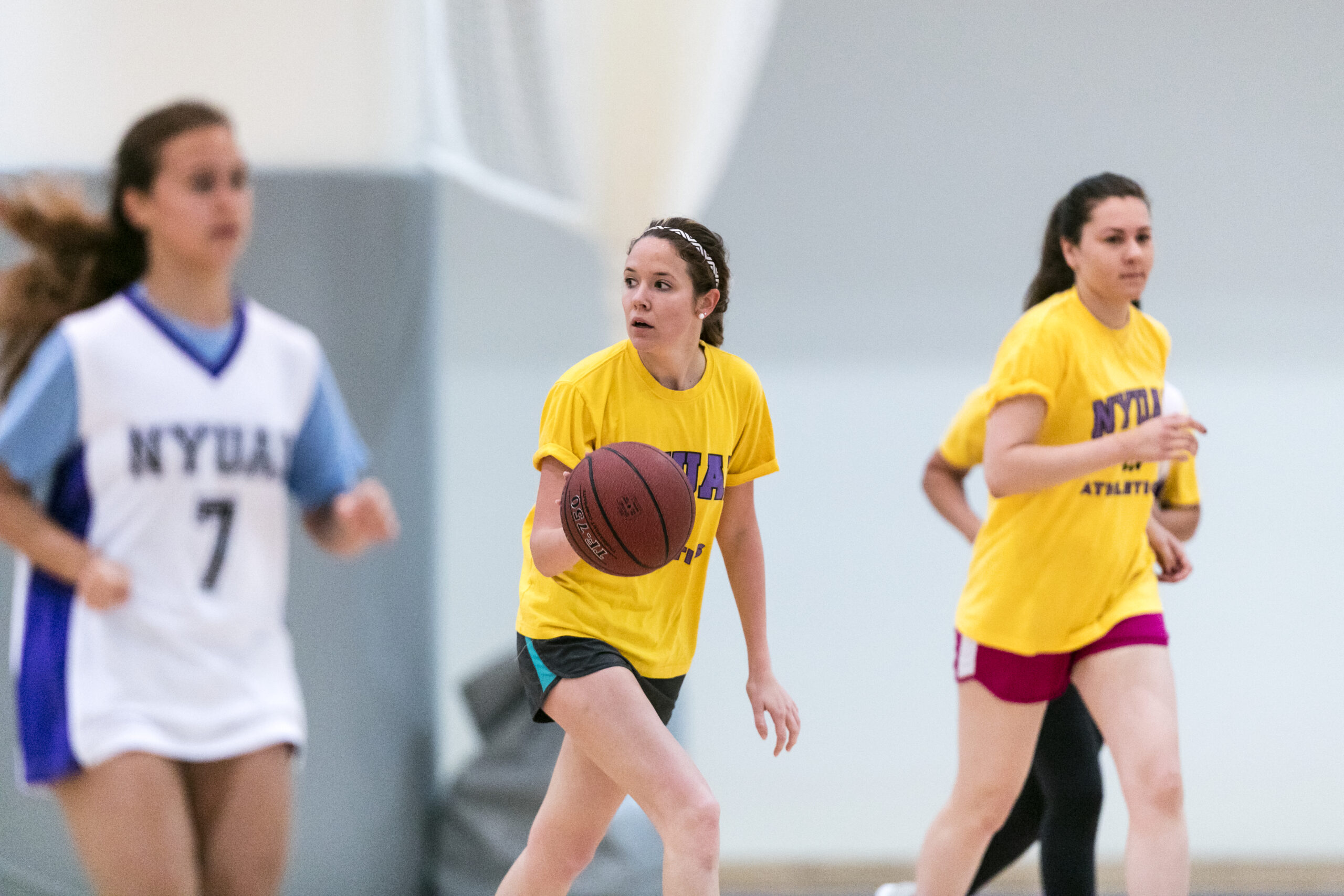 Female basketball players wearing NYU-branded uniforms during a game with one player dribbling the ball.