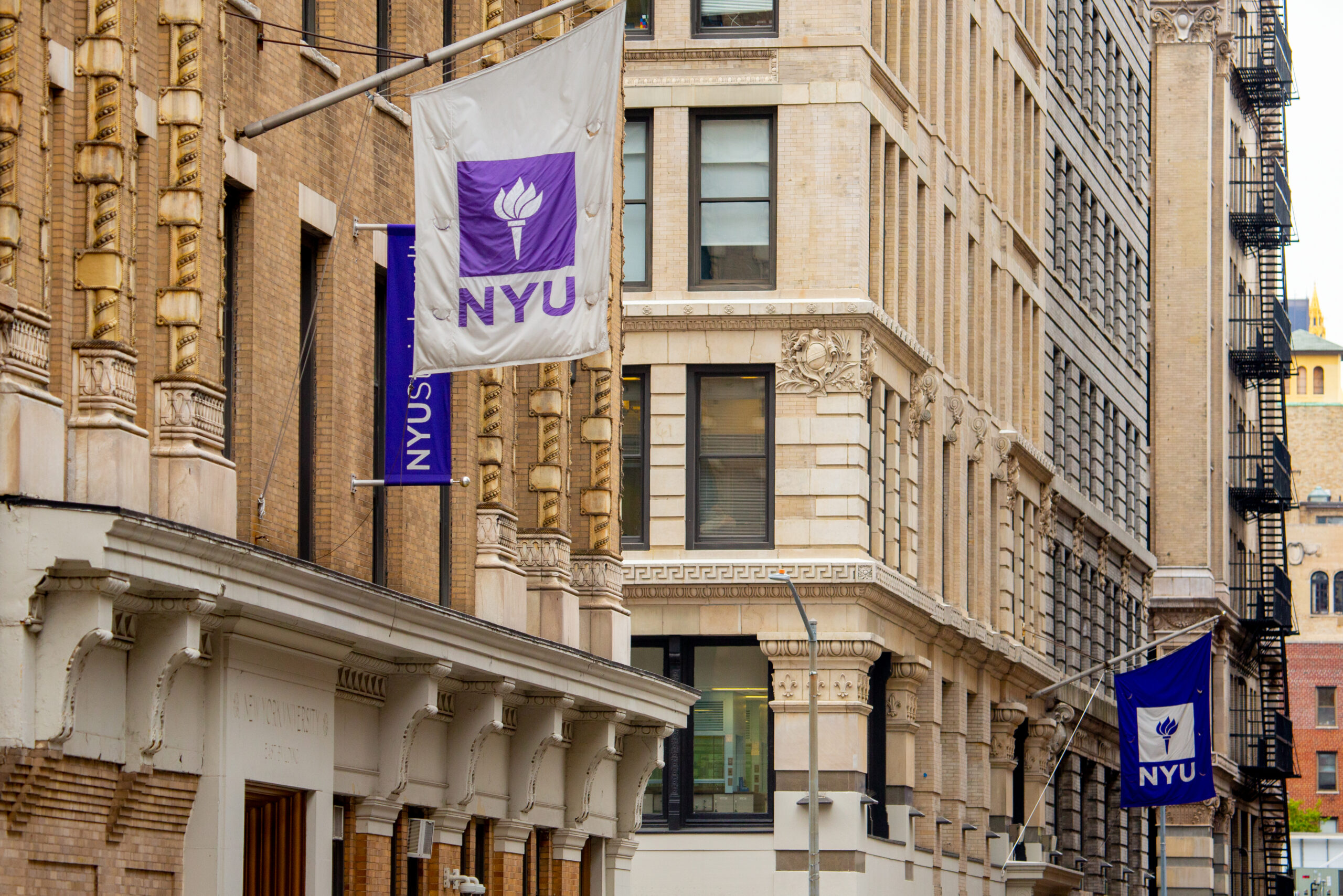 NYU flags hanging on the sides of historic brick buildings on a New York City street.