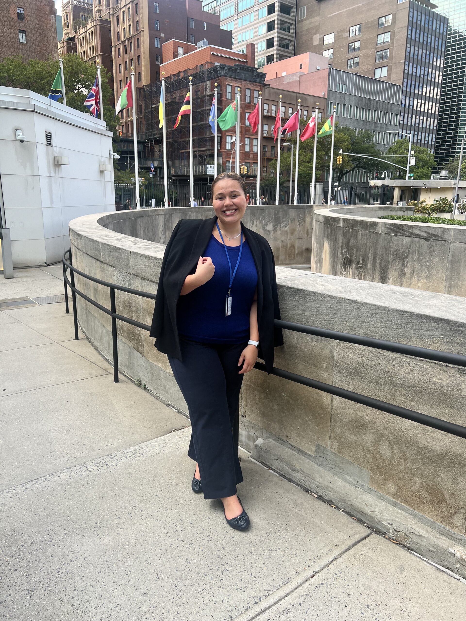 Yulianna Acuna stands outdoors in a formal outfit, wearing a blue top, black slacks, and a black blazer draped over their shoulders. Behind them are international flags.