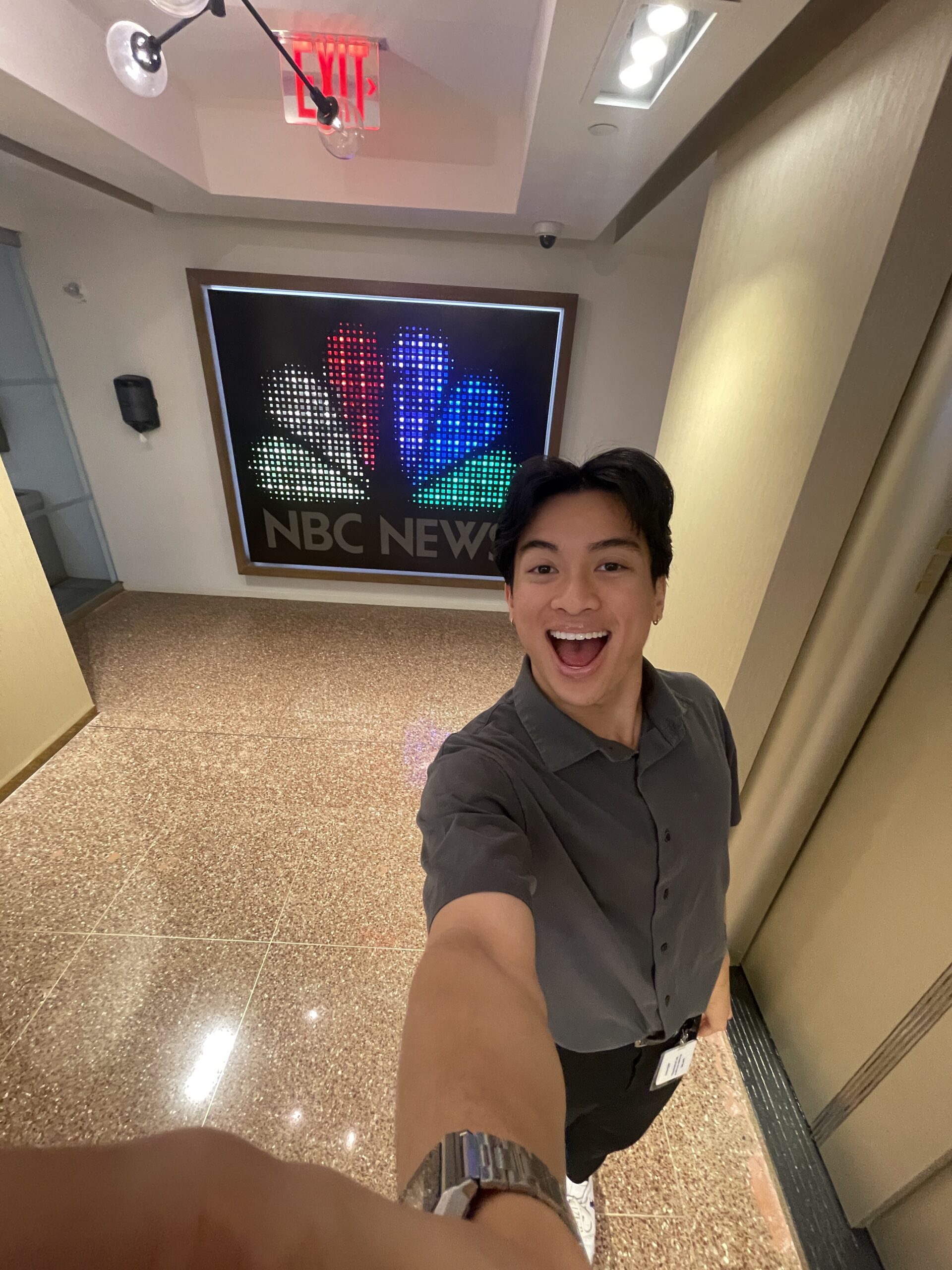 Jake Tran taking a selfie in a hallway with the NBC News logo prominently displayed on a wall behind them. They are wearing a gray button-up shirt and a lanyard, with polished flooring and an exit sign visible above.