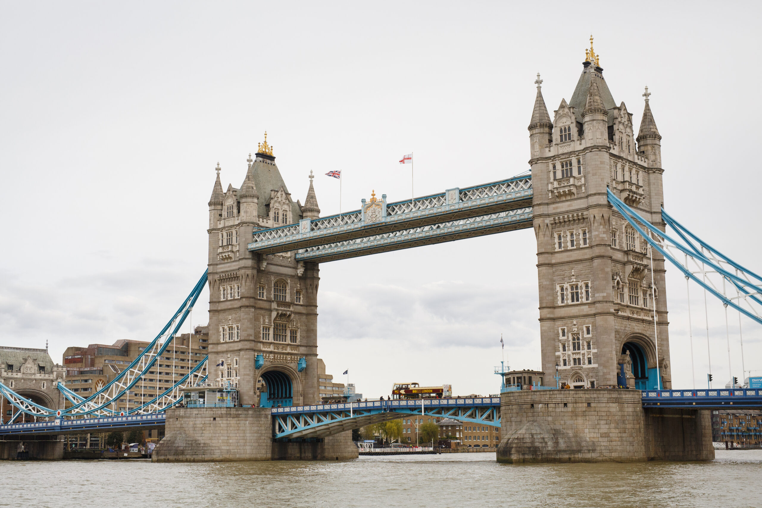 A view of London's iconic Tower Bridge spanning the River Thames with a red double-decker tour bus crossing the bridge.