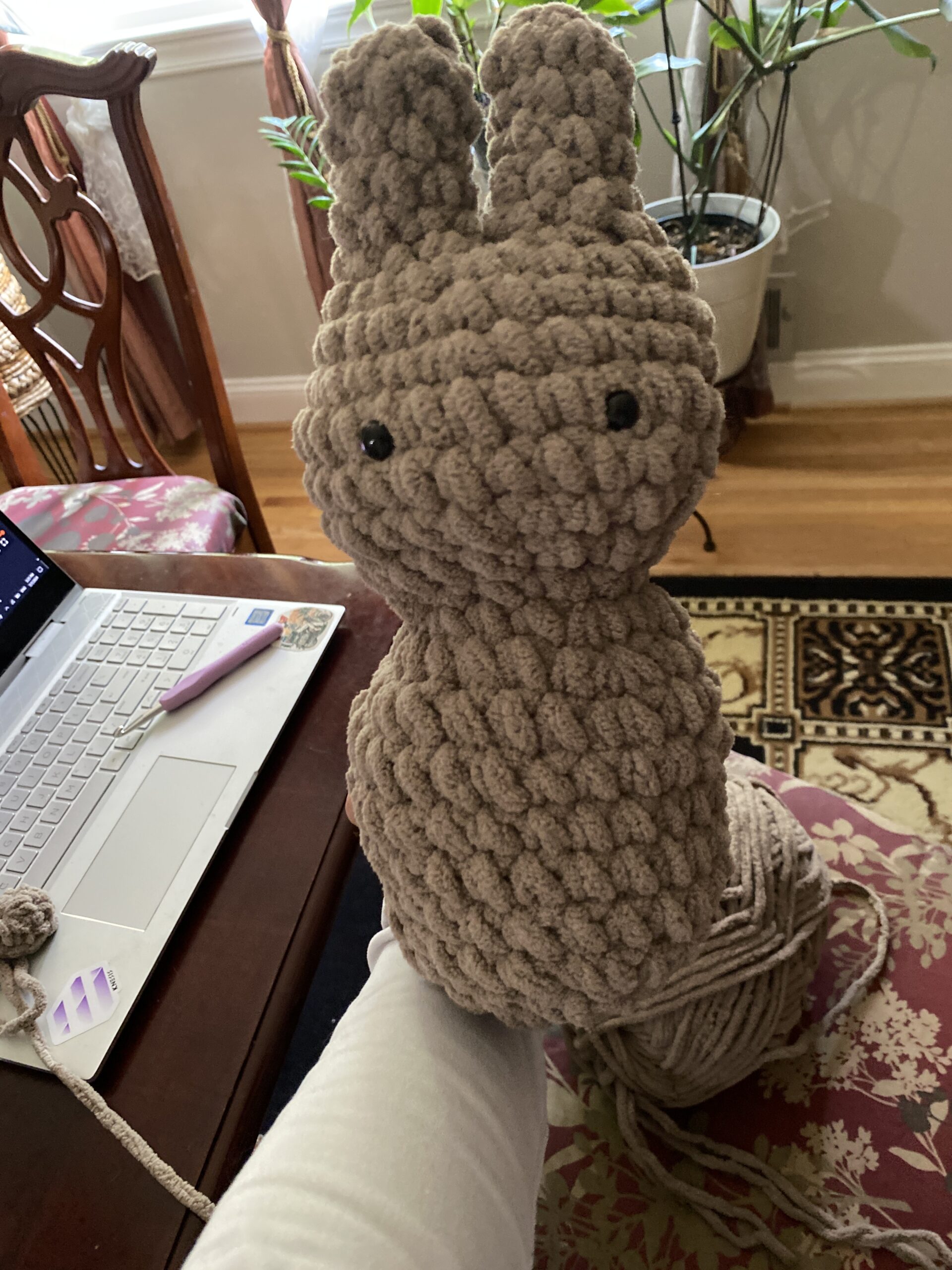 A hand holding a partially completed, plush crocheted bunny made of thick, textured beige yarn, with black safety eyes, in a cozy indoor setting with a laptop and yarn in the background.