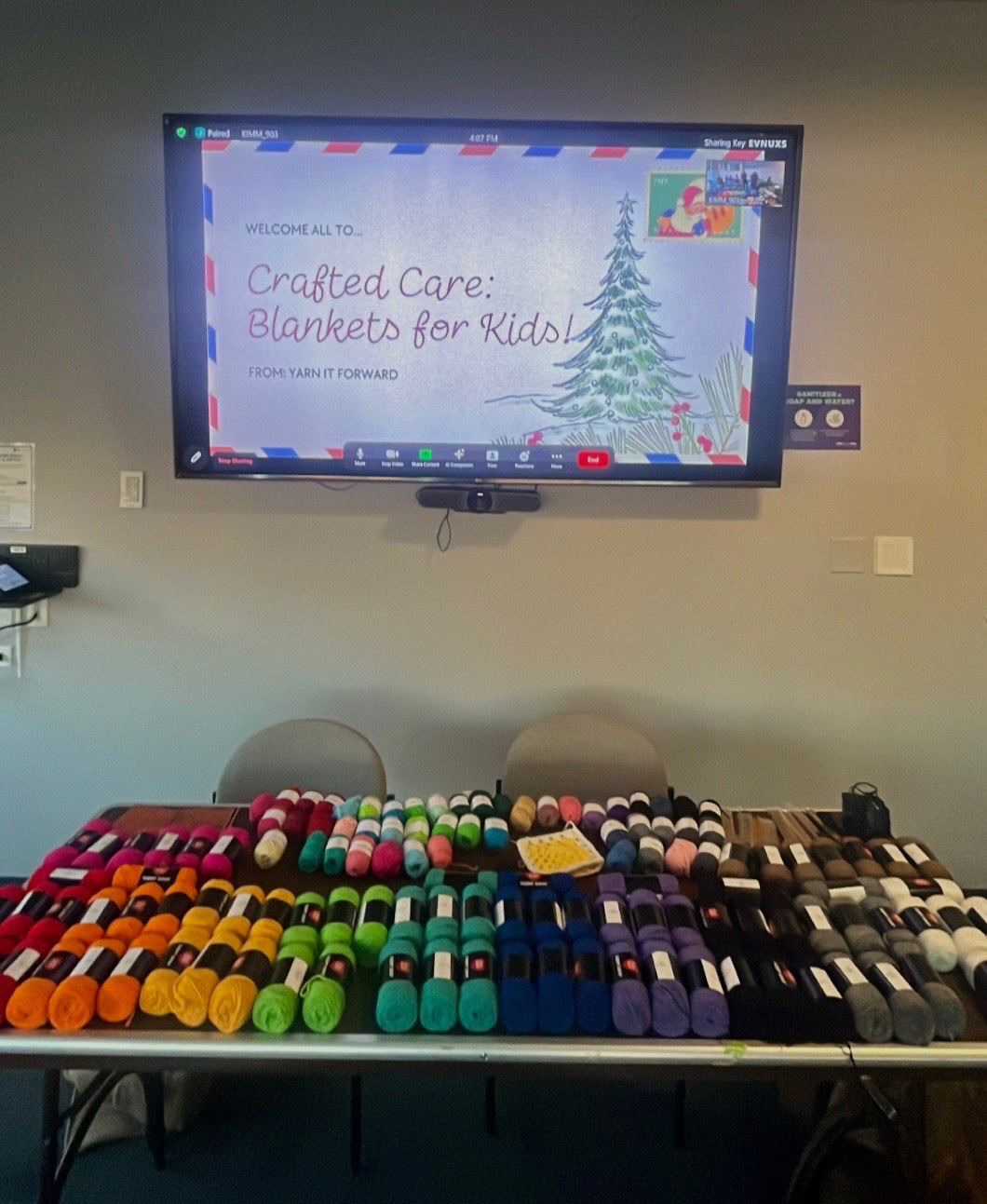 A table filled with colorful skeins of yarn is set up in front of a screen displaying a virtual event titled