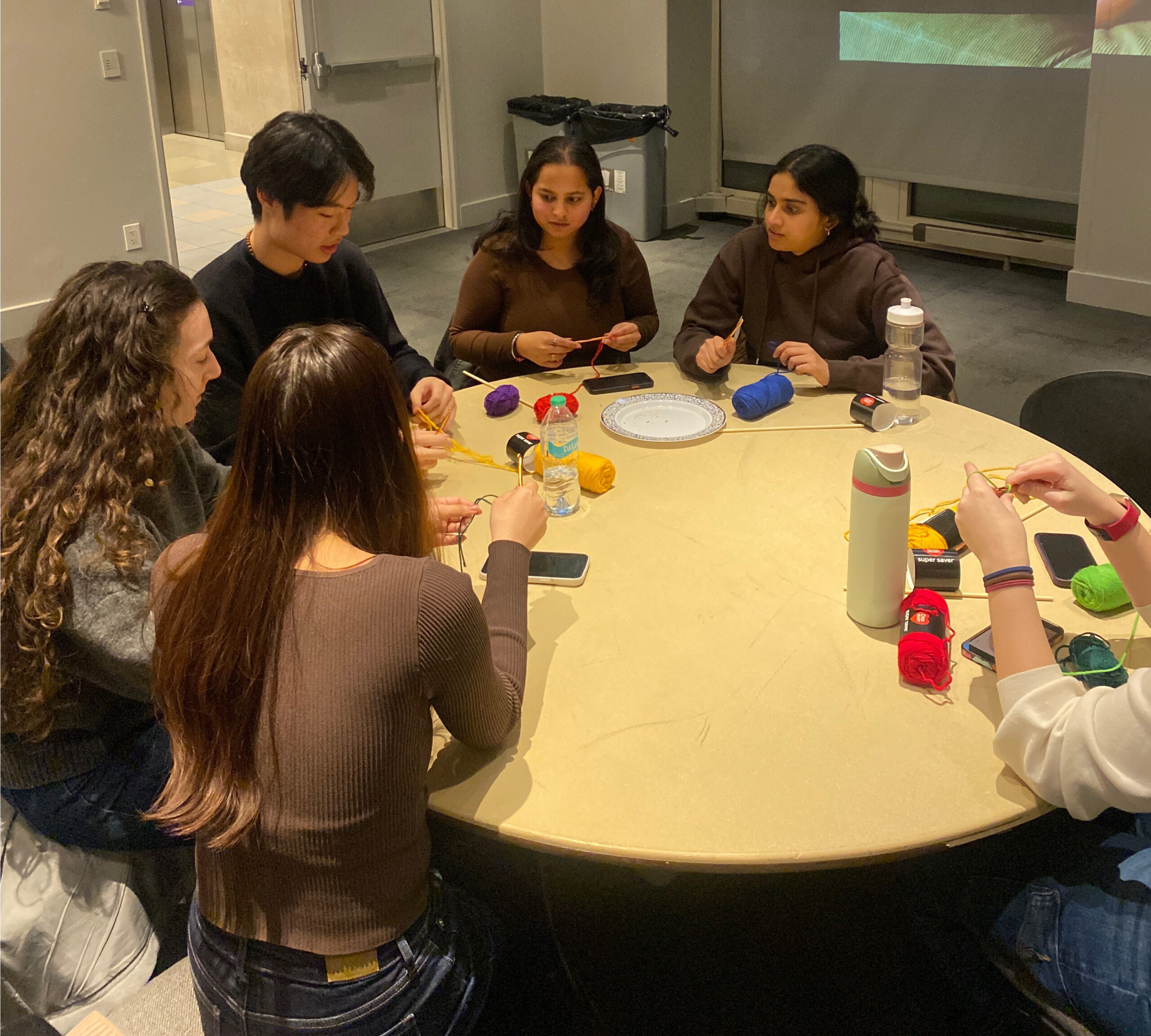 A group of people sitting around a table, engaged in crocheting with colorful yarn, as part of a crafting event in a communal setting.