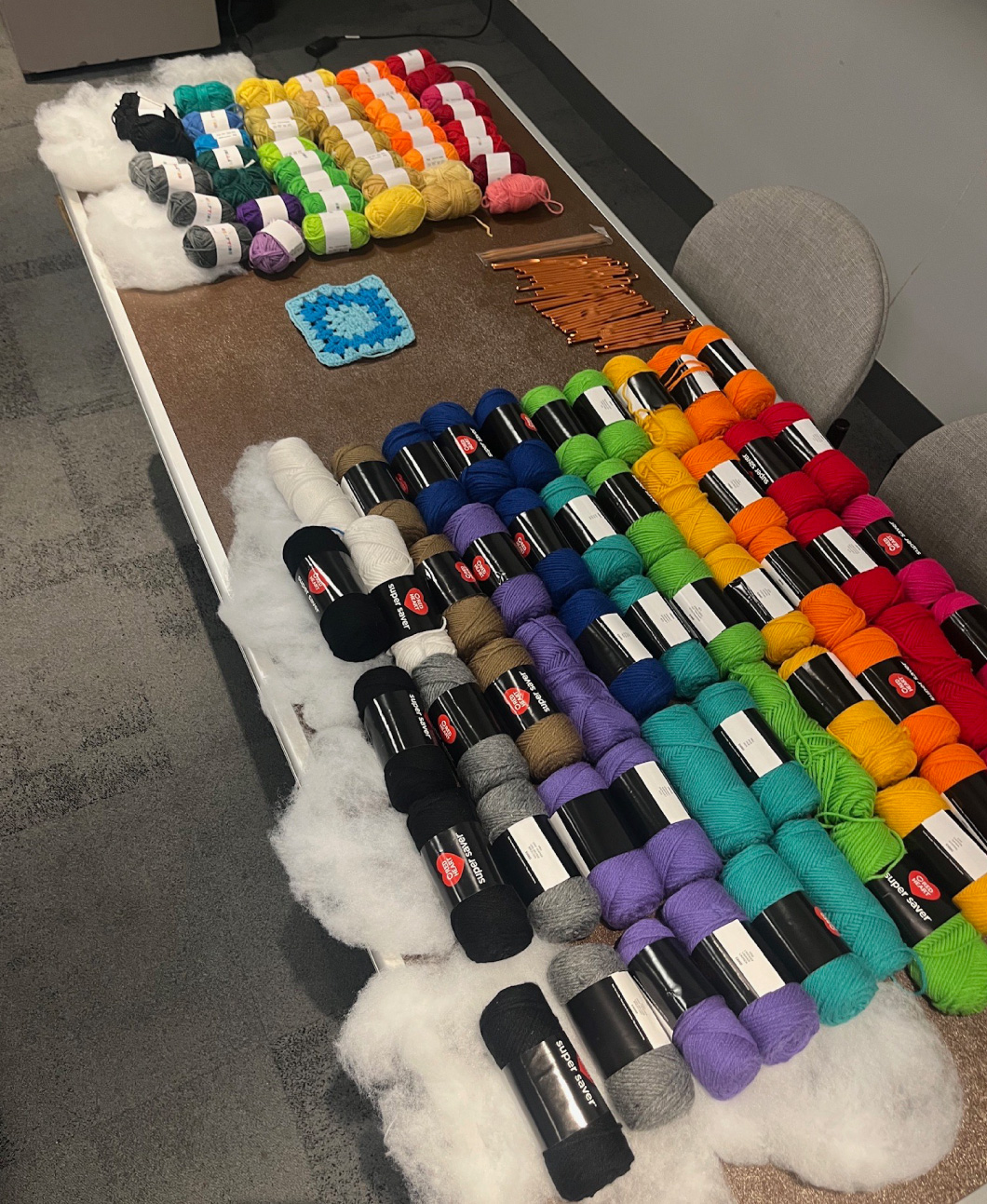 A table covered with colorful skeins of yarn, crochet hooks, and a single blue granny square, set up for a crafting event, with fluffy batting along the edges for decoration.