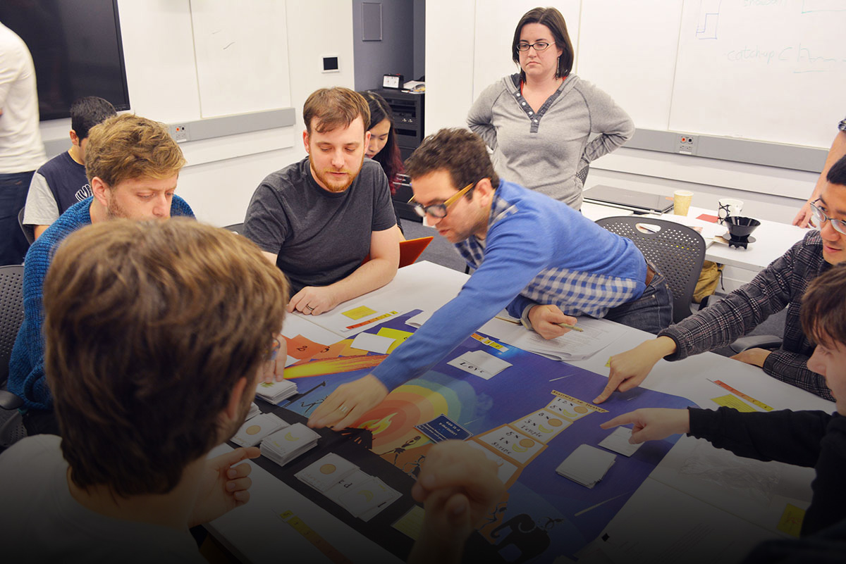 students gather around a board game on a table to play test