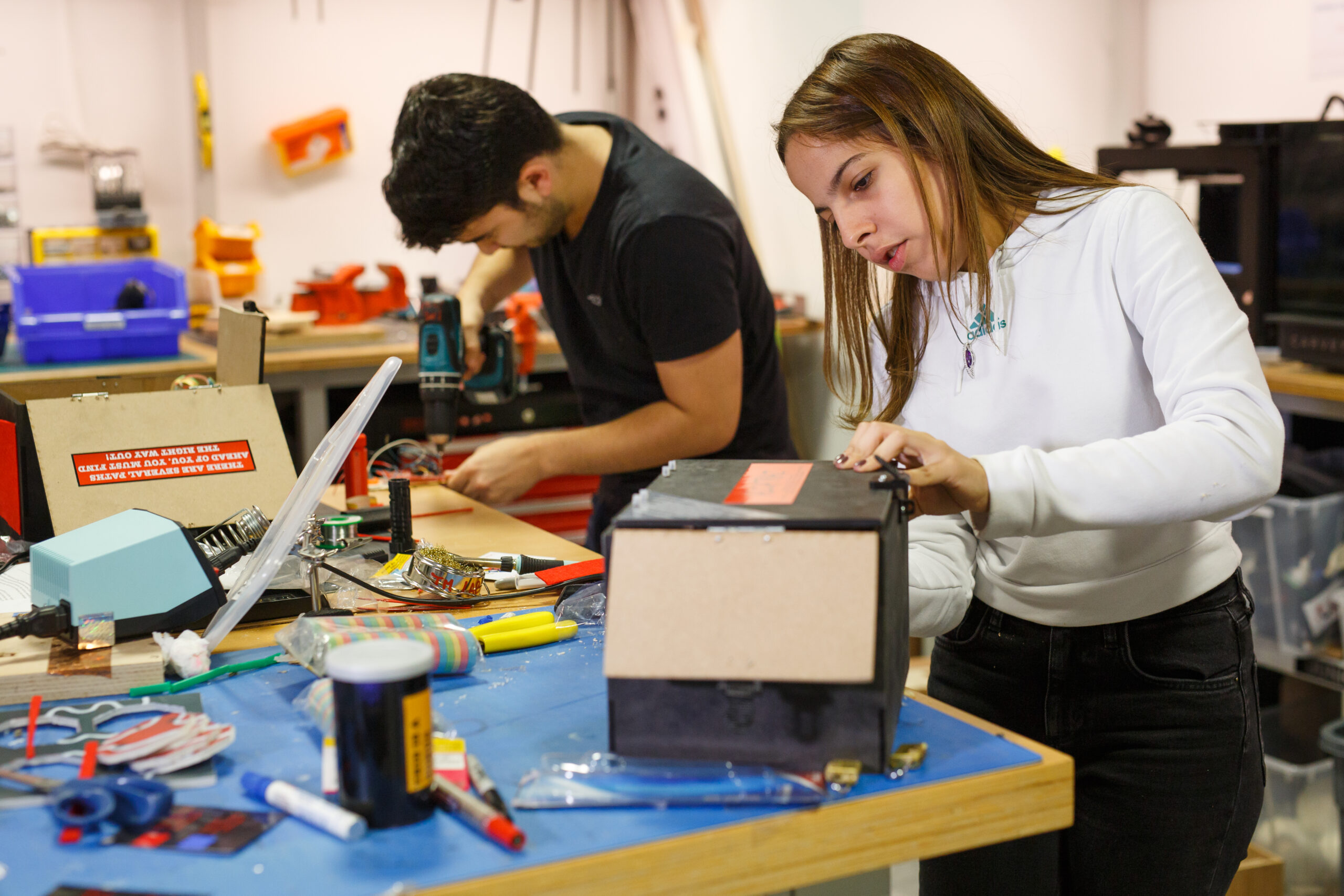 Two students working on projects in a workshop, with one using a power drill while the other assembles a black box with red labels.