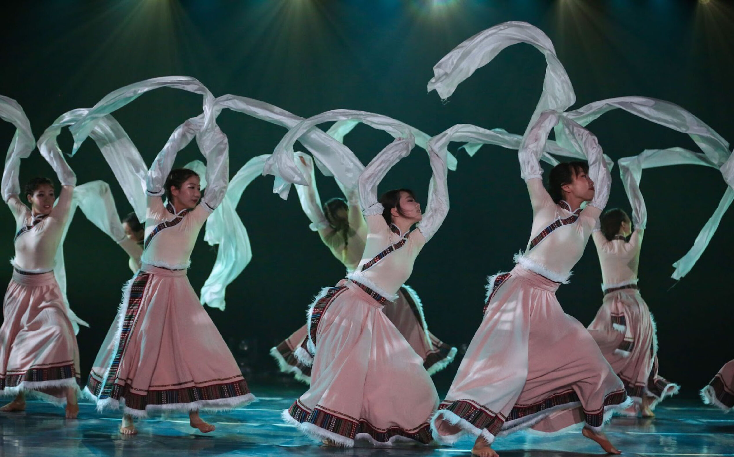Dancers performing on stage in pink long skirts and moving large white scarves.