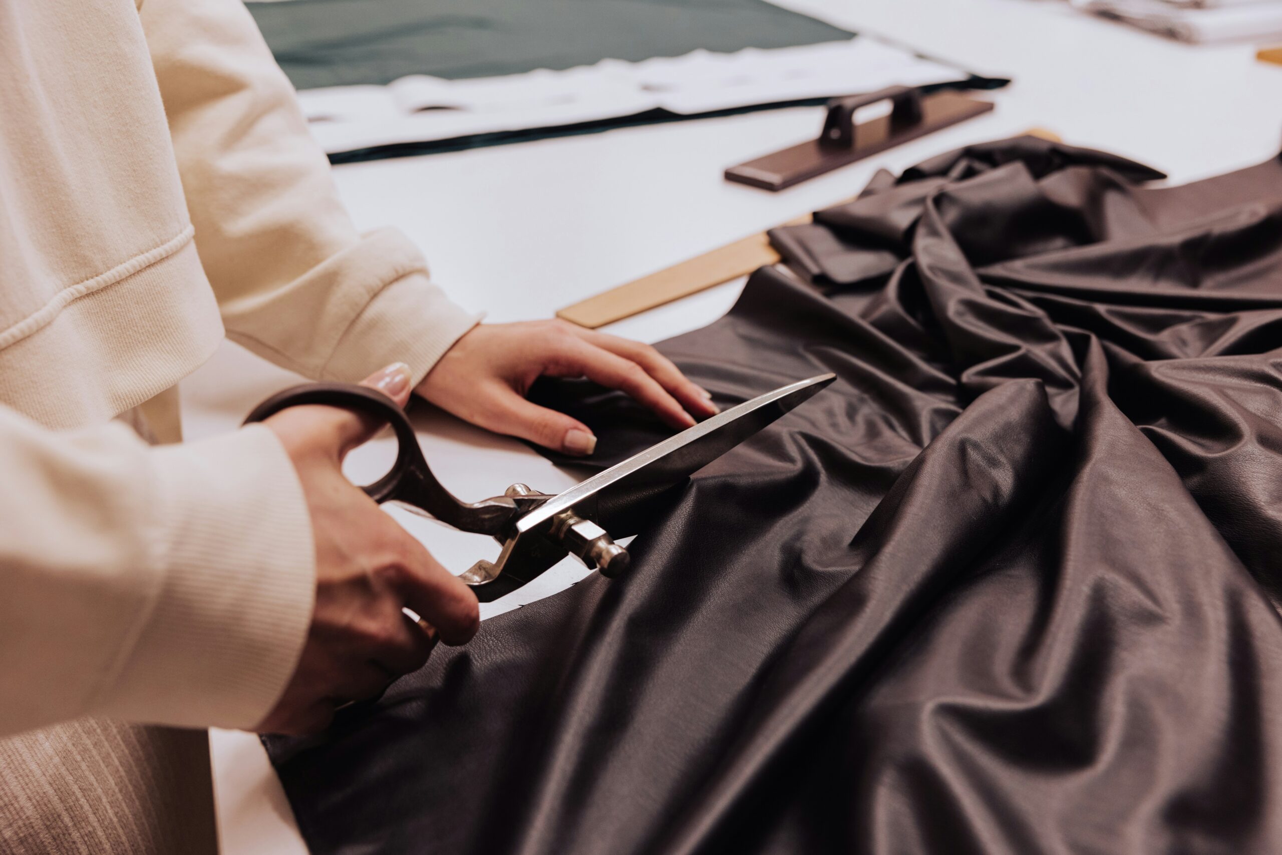 A person carefully cuts a piece of dark fabric with large tailor’s shears on a worktable, preparing materials for a sewing or fashion design project.