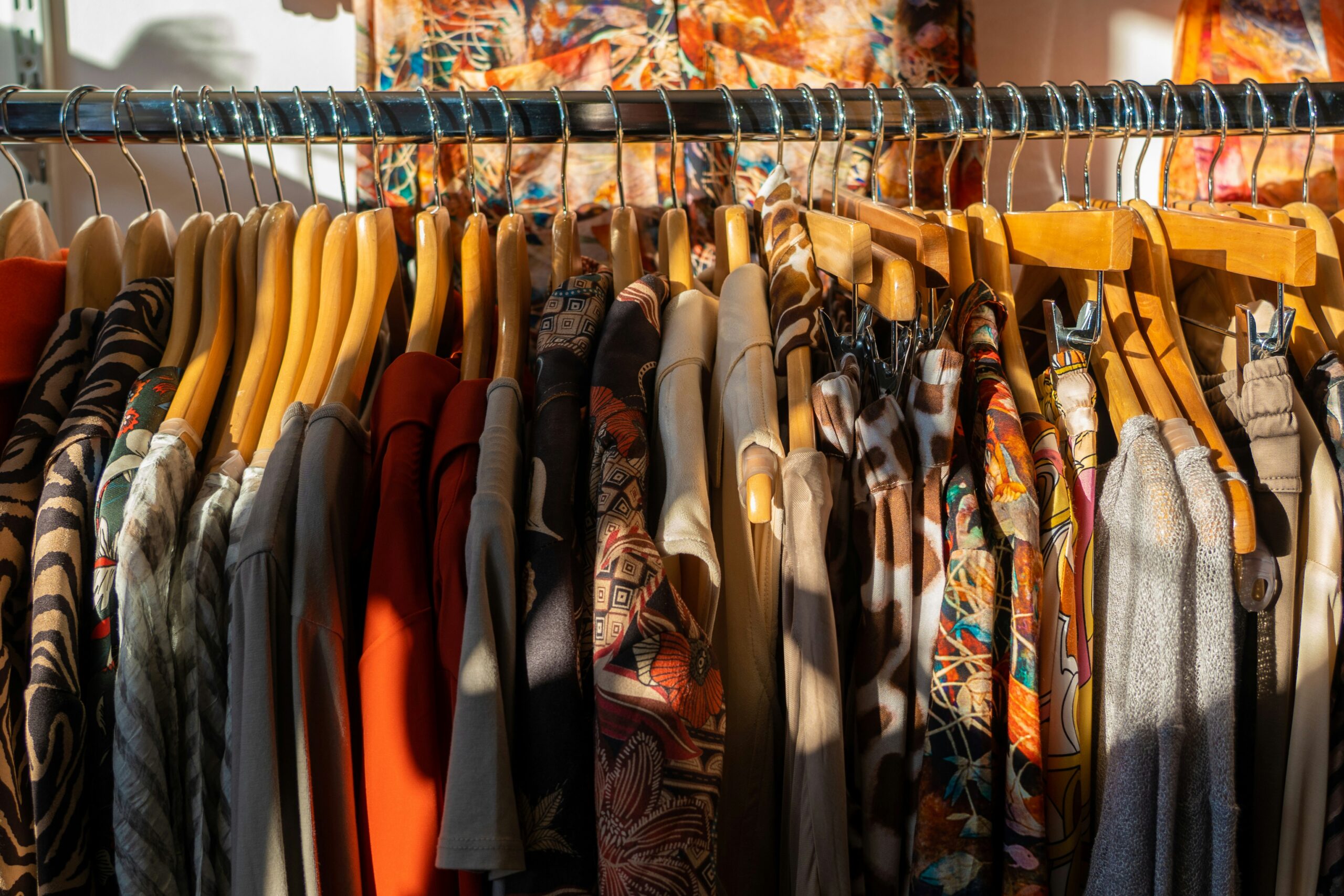 Colorful, bold clothing in a variety of textures and patterns fills a clothing rack warmly lit by sunlight.