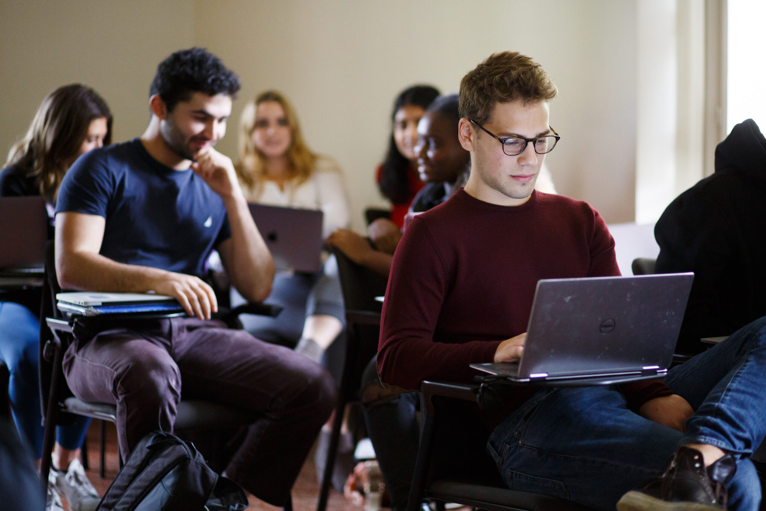 A student is seated working on their laptop while a group of students holds a discussion behind them.