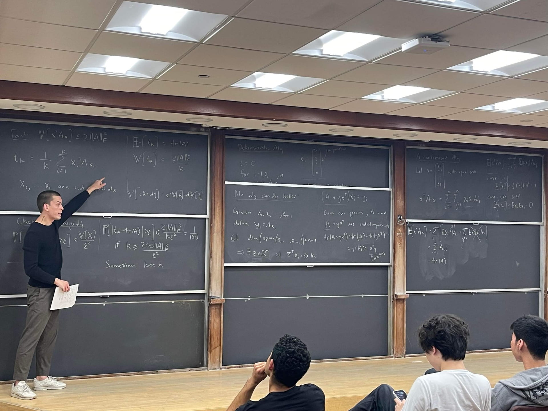 A student standing and presenting at a board with numerous math equations to seated students