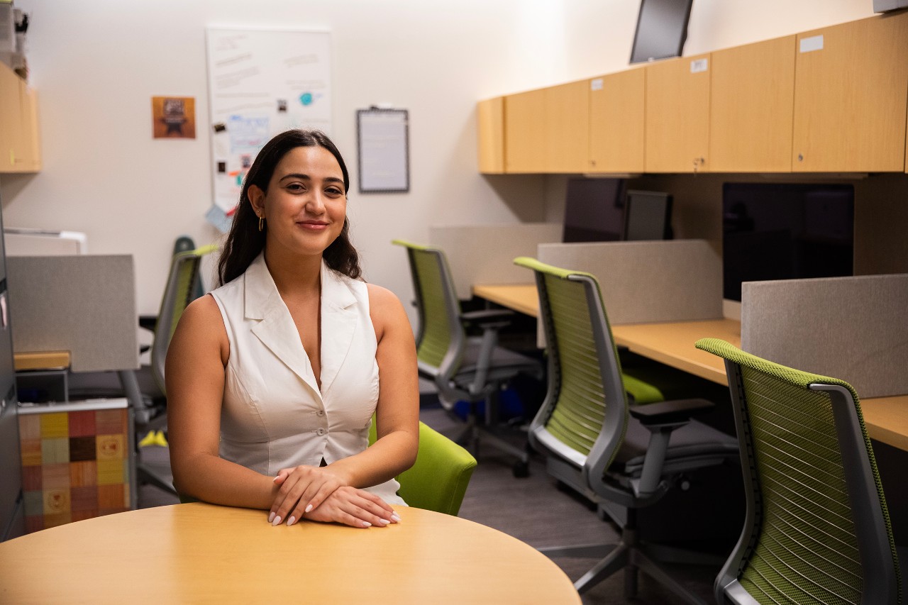 Portrait of Jida Haddad, sitting with hands clasped at a table with a row of cubby desks in the background.
