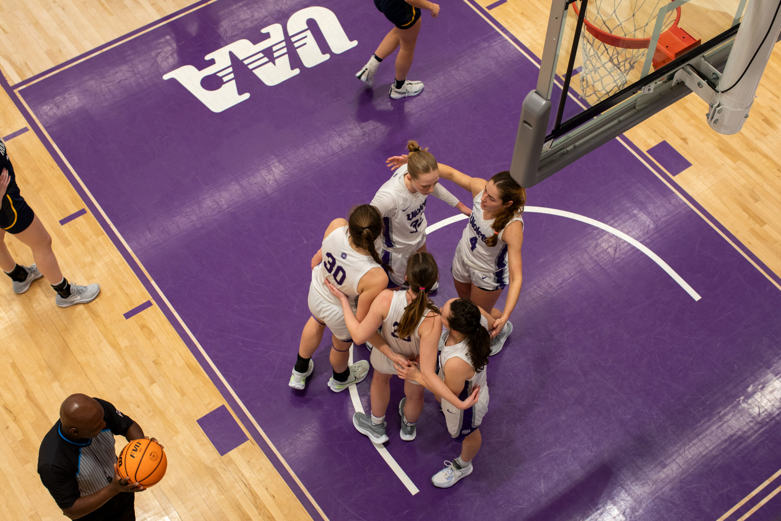 NYU women’s basketball players huddle together under the basket. The referee holds the ball nearby as they prepare to resume the game.