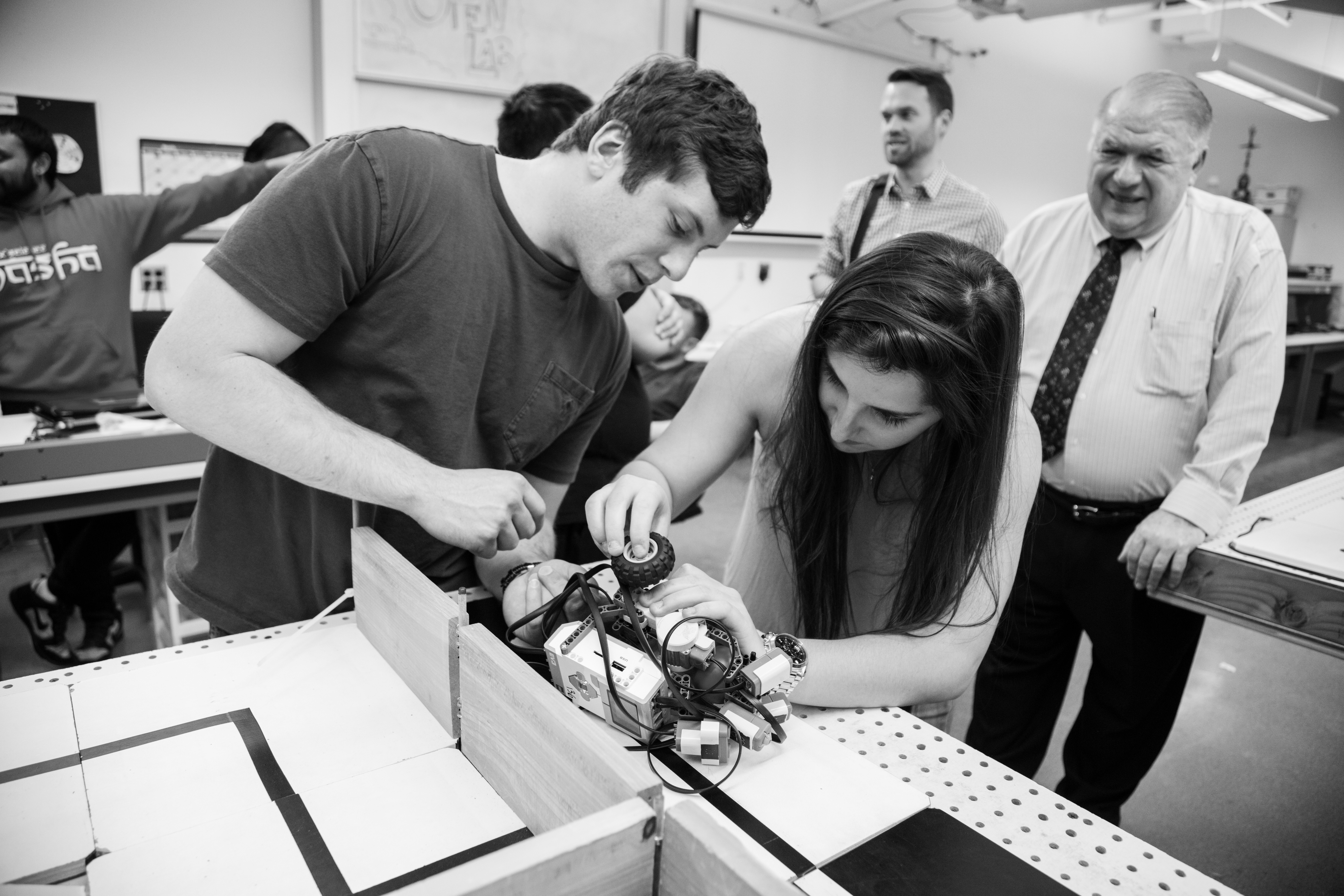 Students work together on assembling and testing a small robot in a classroom or lab setting, while instructors and peers observe in the background.