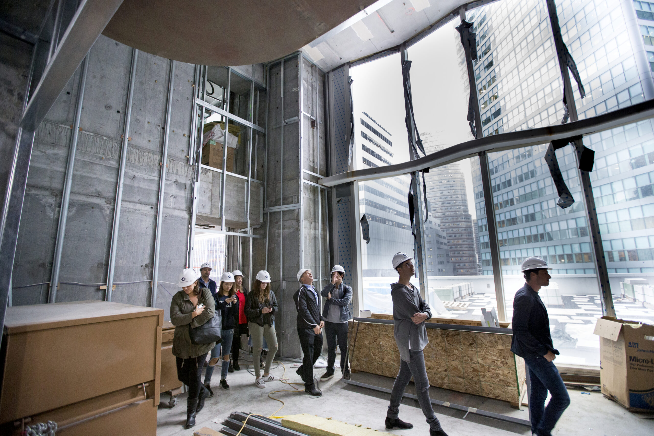 A group of students wearing hard hats tour a building under construction with large windows overlooking city skyscrapers.