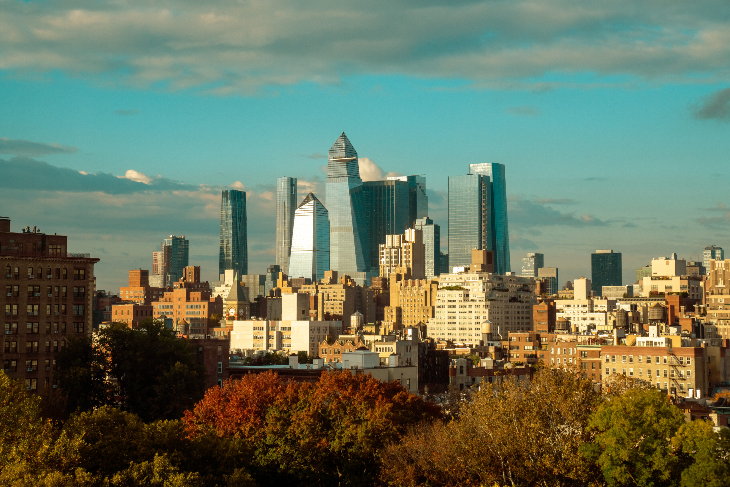 New York City skyline with tall glass buildings and colorful autumn trees in the foreground.