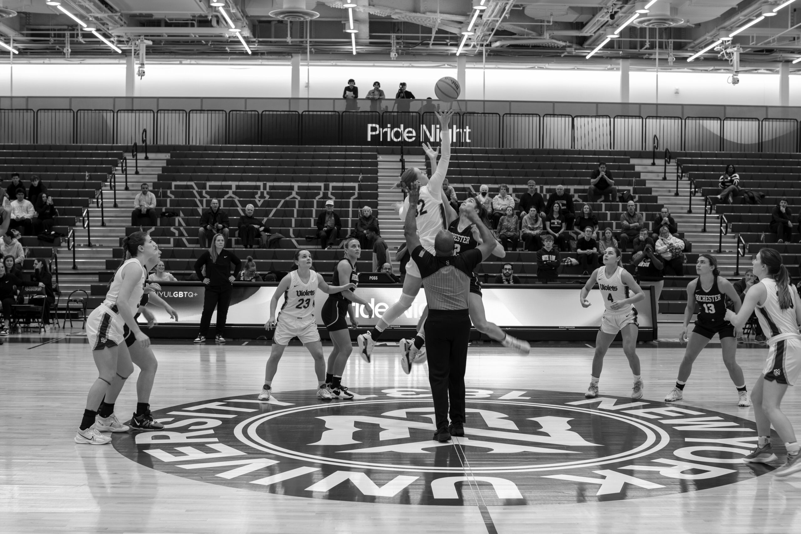 A black-and-white photo of an NYU women’s basketball game tip-off, with players from NYU Violets and Rochester jumping for the ball under a banner that reads “Pride Night.”