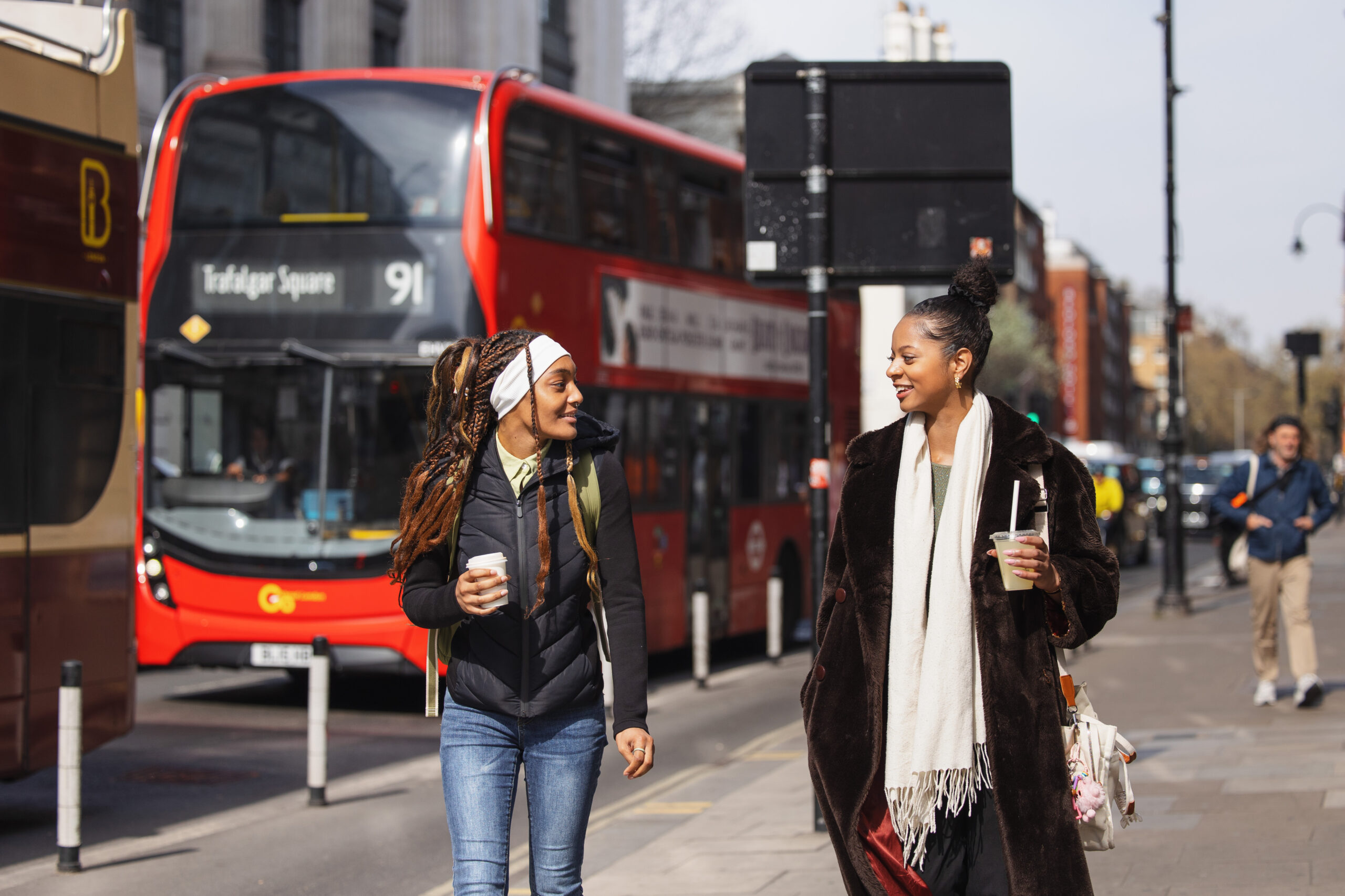 Two women walk and chat on a London street as a red double-decker bus makes its way to Trafalgar Square behind them.