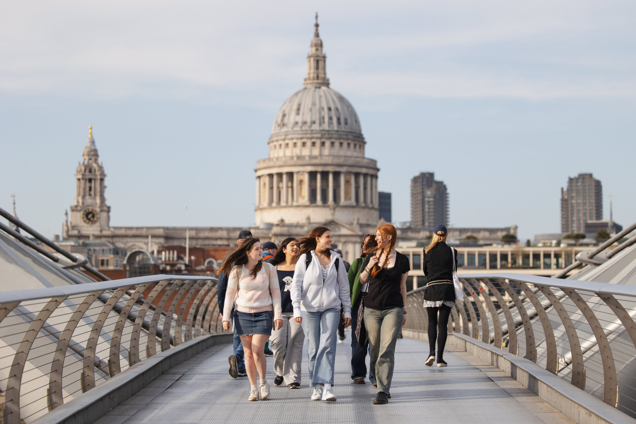 A group of people walk across the Millennium Bridge in London on a clear day, with St. Paul’s Cathedral prominently visible in the background.