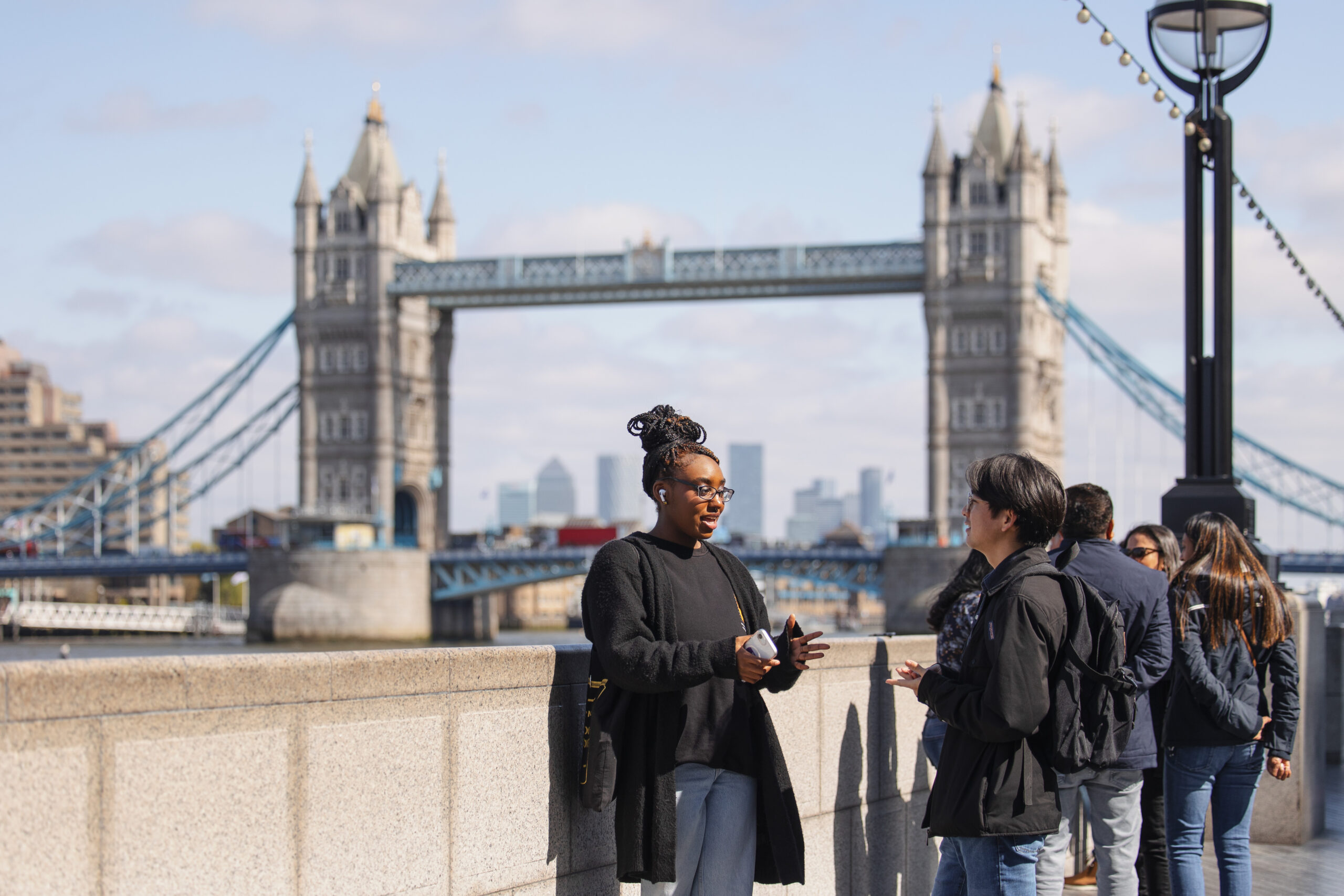 Two people talk along the River Thames on a sunny day, Tower Bridge spreading across the sky behind them.