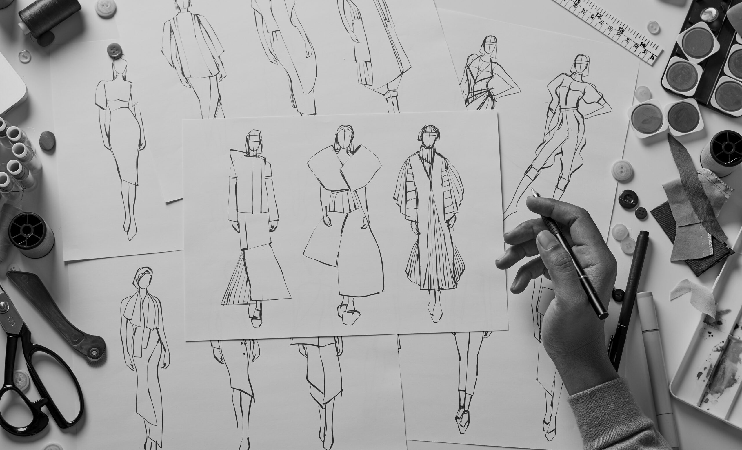 A designer’s hand holds a pen over fashion sketches of models in various outfits, surrounded by sewing tools, buttons, fabric swatches, and paints on a workspace.