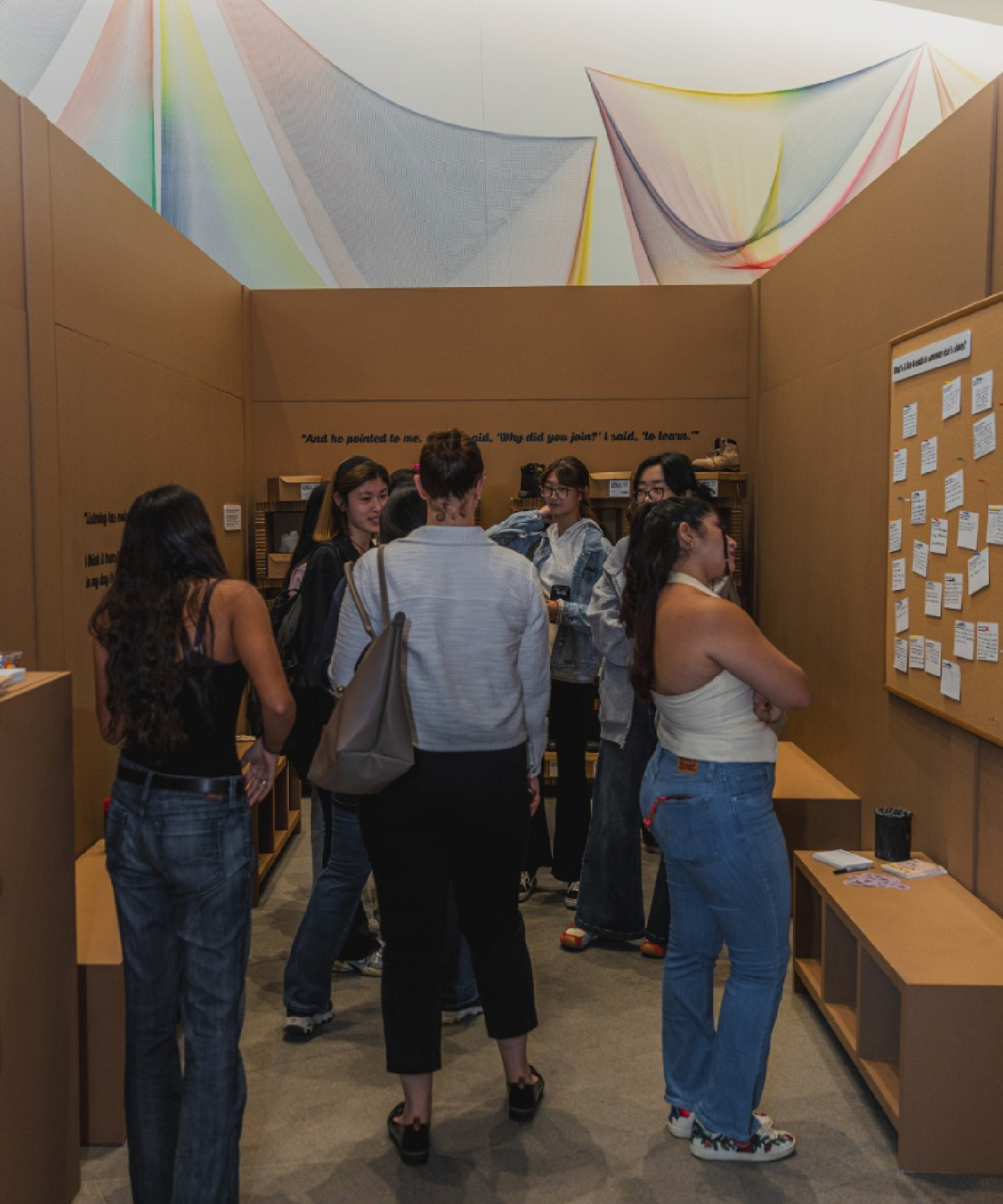 A group of students gather inside the Empathy Museum’s interactive exhibit space, reading and discussing notes pinned on a bulletin board. The walls are lined with text and displays, with colorful fabric art hanging above.