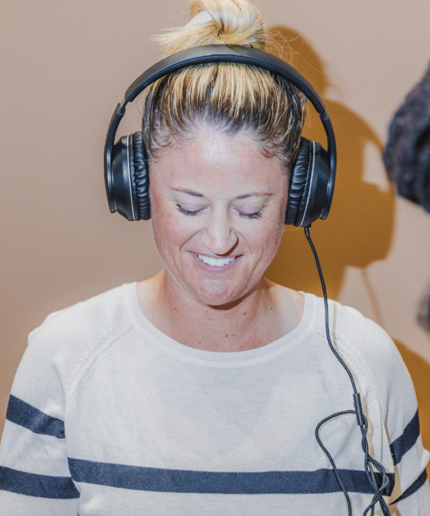 A person wearing headphones smiles while looking down at a phone. They are dressed in a white sweater with navy stripes and appear to be listening to audio.