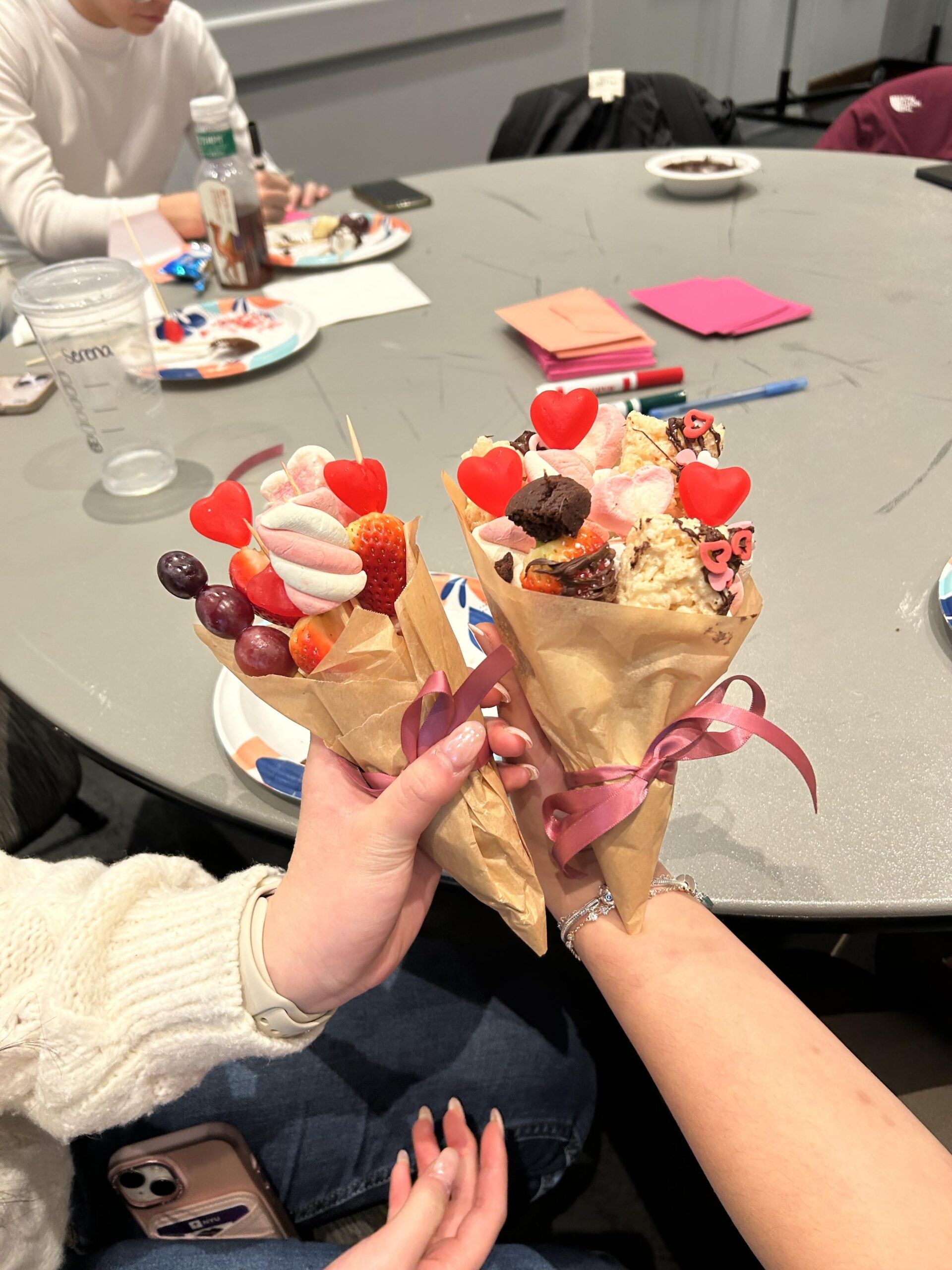 Two students hold up decorative paper cones filled with fruit, marshmallows, cookies, and heart-shaped treats, tied with pink ribbons during an NYU CHEFs for School club event.
