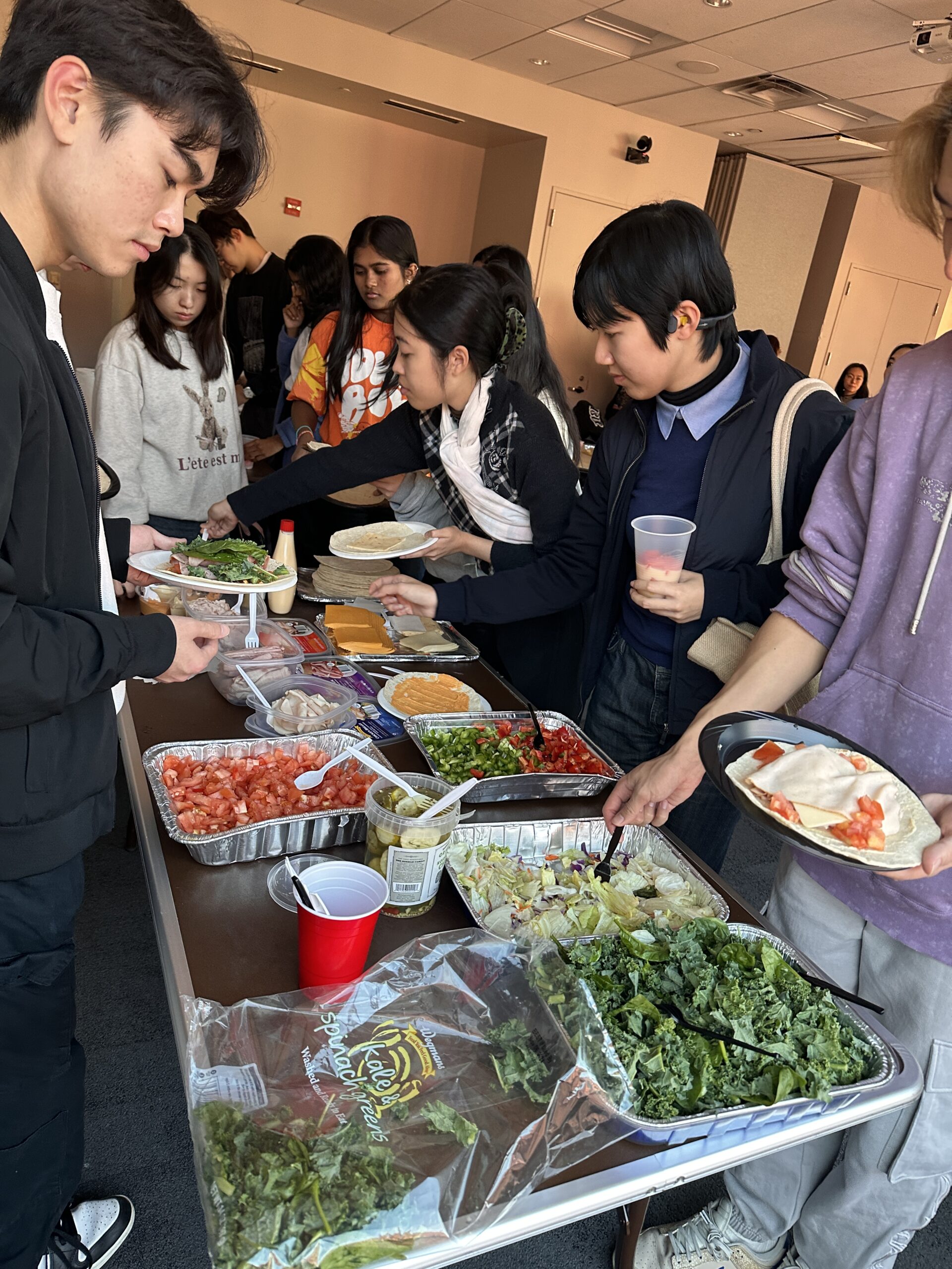 Students line up at a buffet table during an NYU CHEFs for School club event, filling their plates with wraps, vegetables, cheese, and salad.