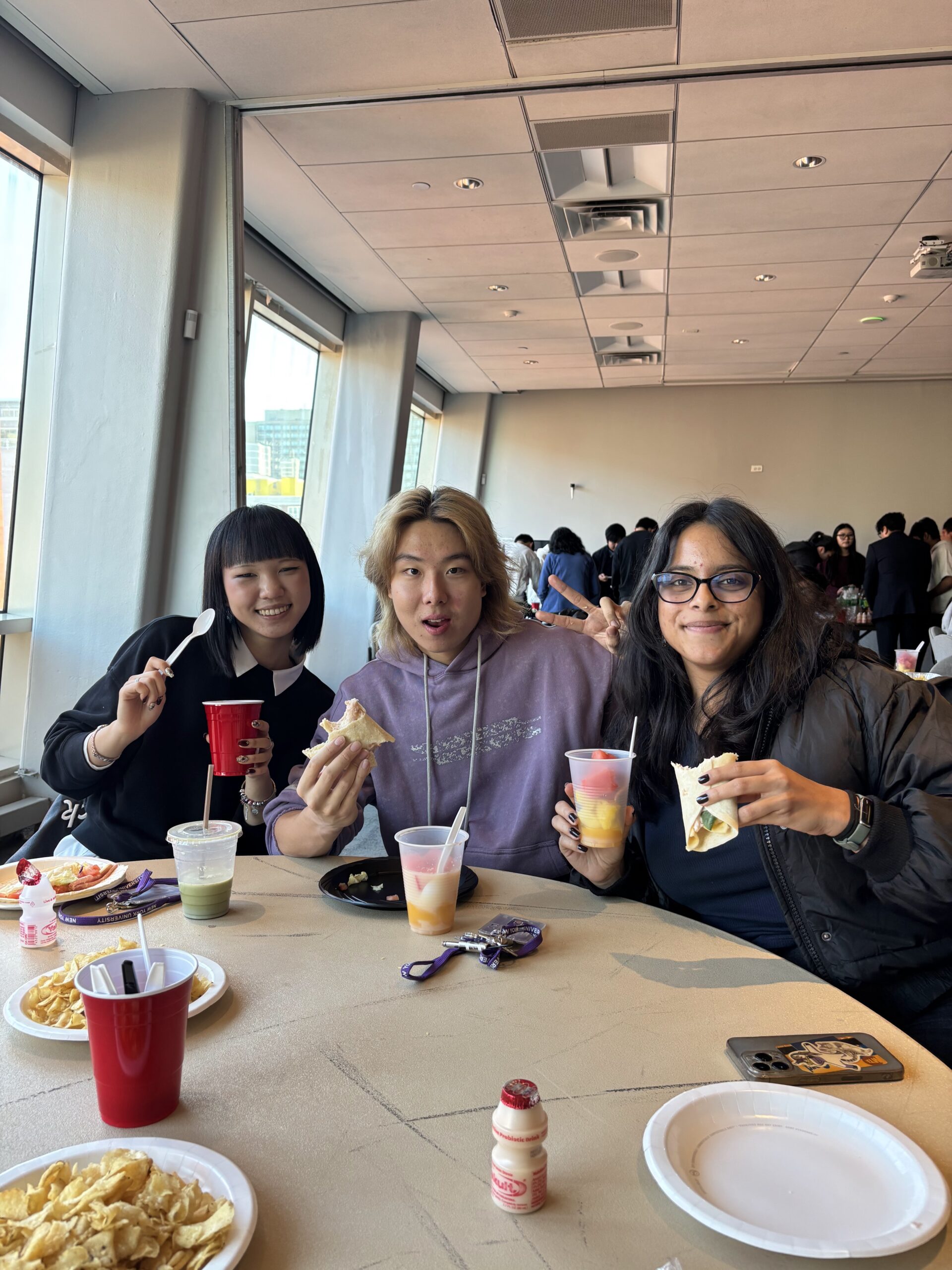 Three students sit together at a table during an NYU CHEFs for School club event, smiling and holding wraps and fruit cups, with snacks and drinks spread out in front of them.