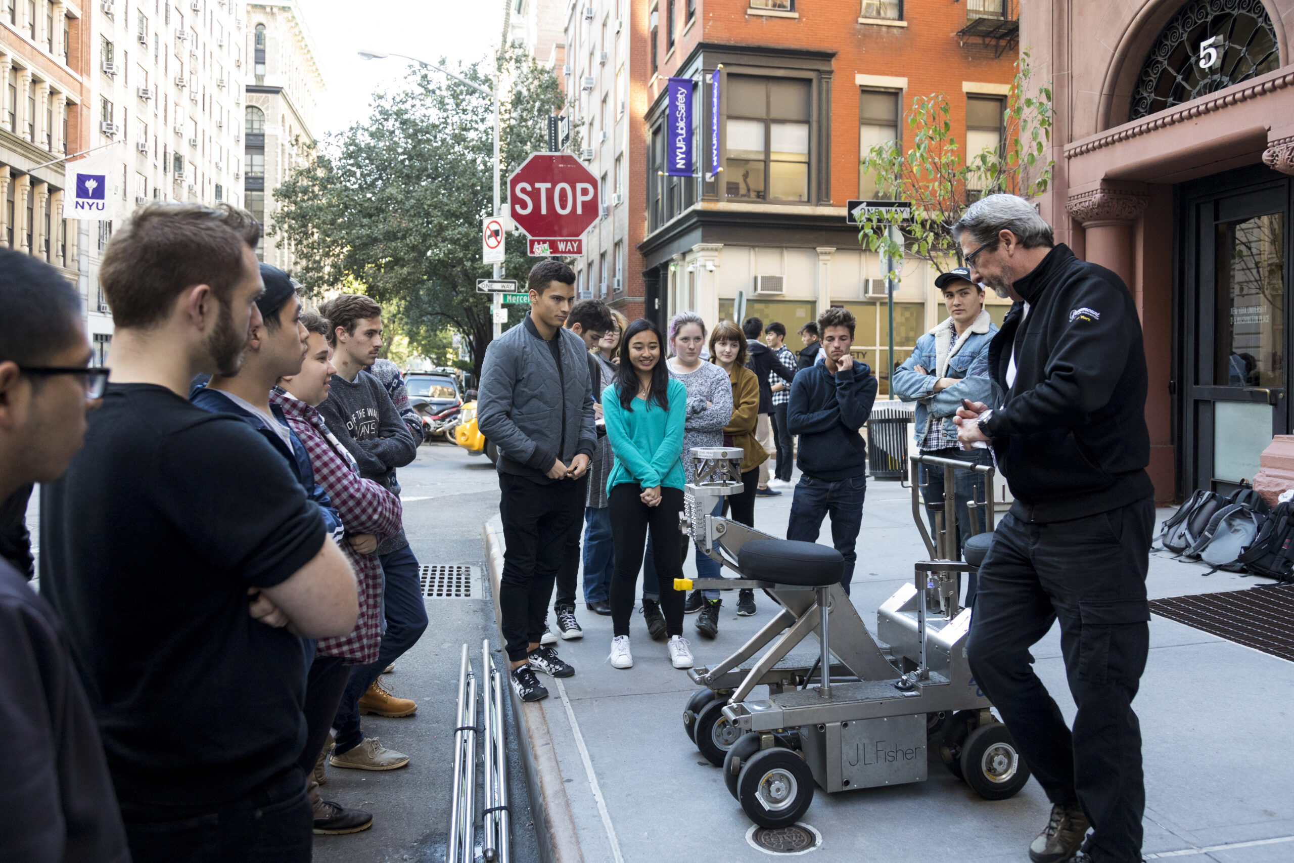 A large group of students and a professor stand together on a New York City street corner, gathered around a large piece of filming equipment.