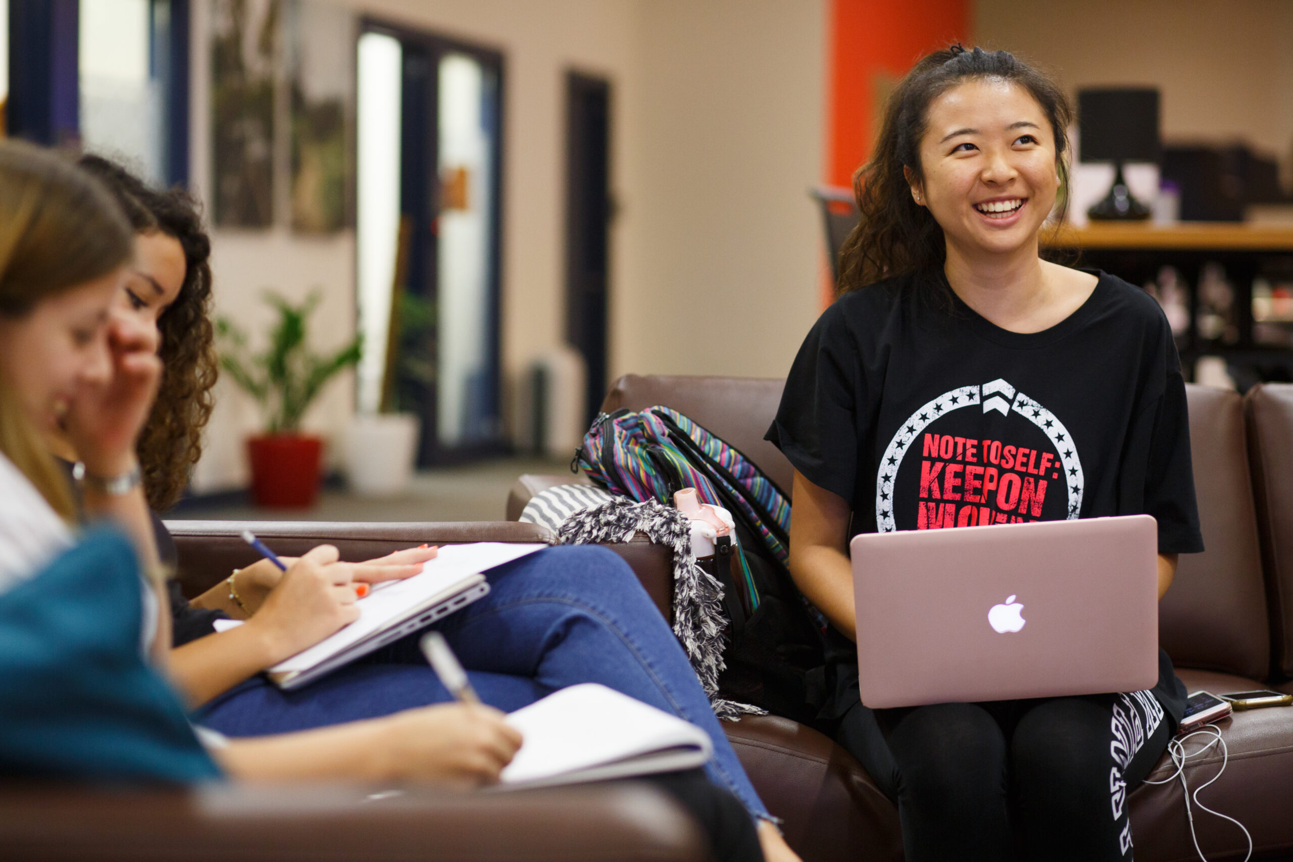 Three students sit together on couches, smiling and taking notes as one works on a laptop during a casual study session.