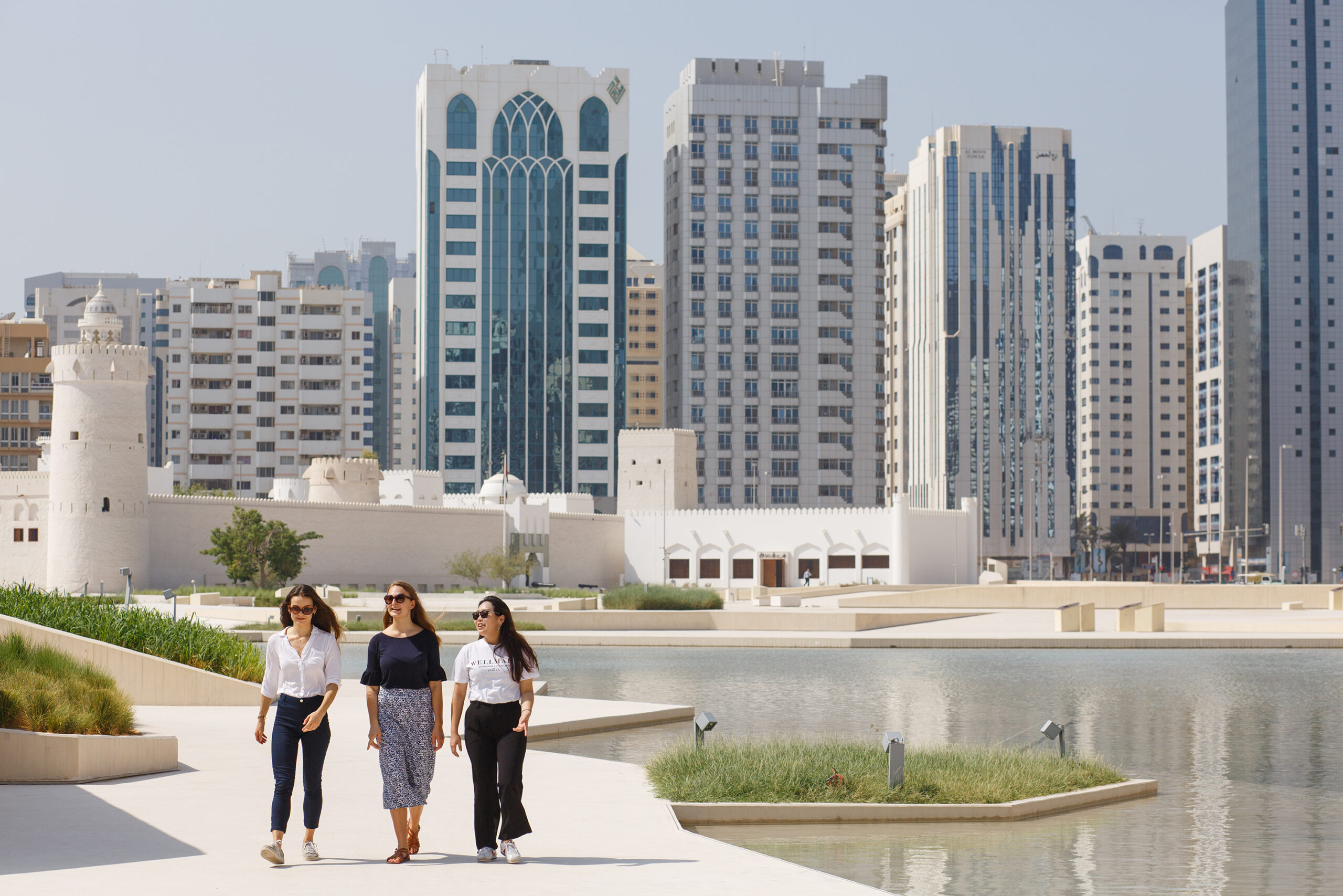 Three students walk together by a reflecting pool in Abu Dhabi, with modern high-rise buildings and traditional architecture visible in the background.
