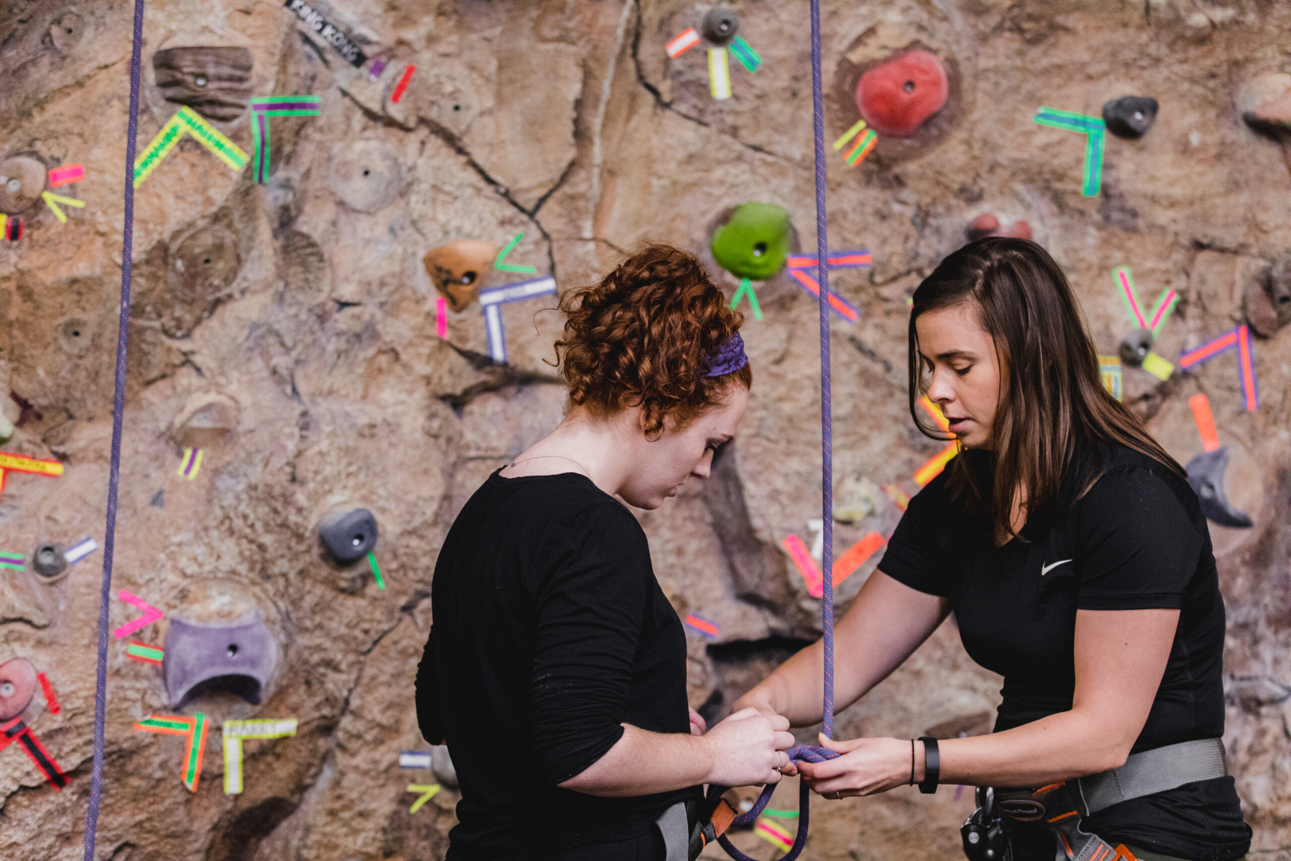 Two people prepare for indoor rock climbing as one helps the other secure a harness rope against a climbing wall marked with colorful tape and holds.