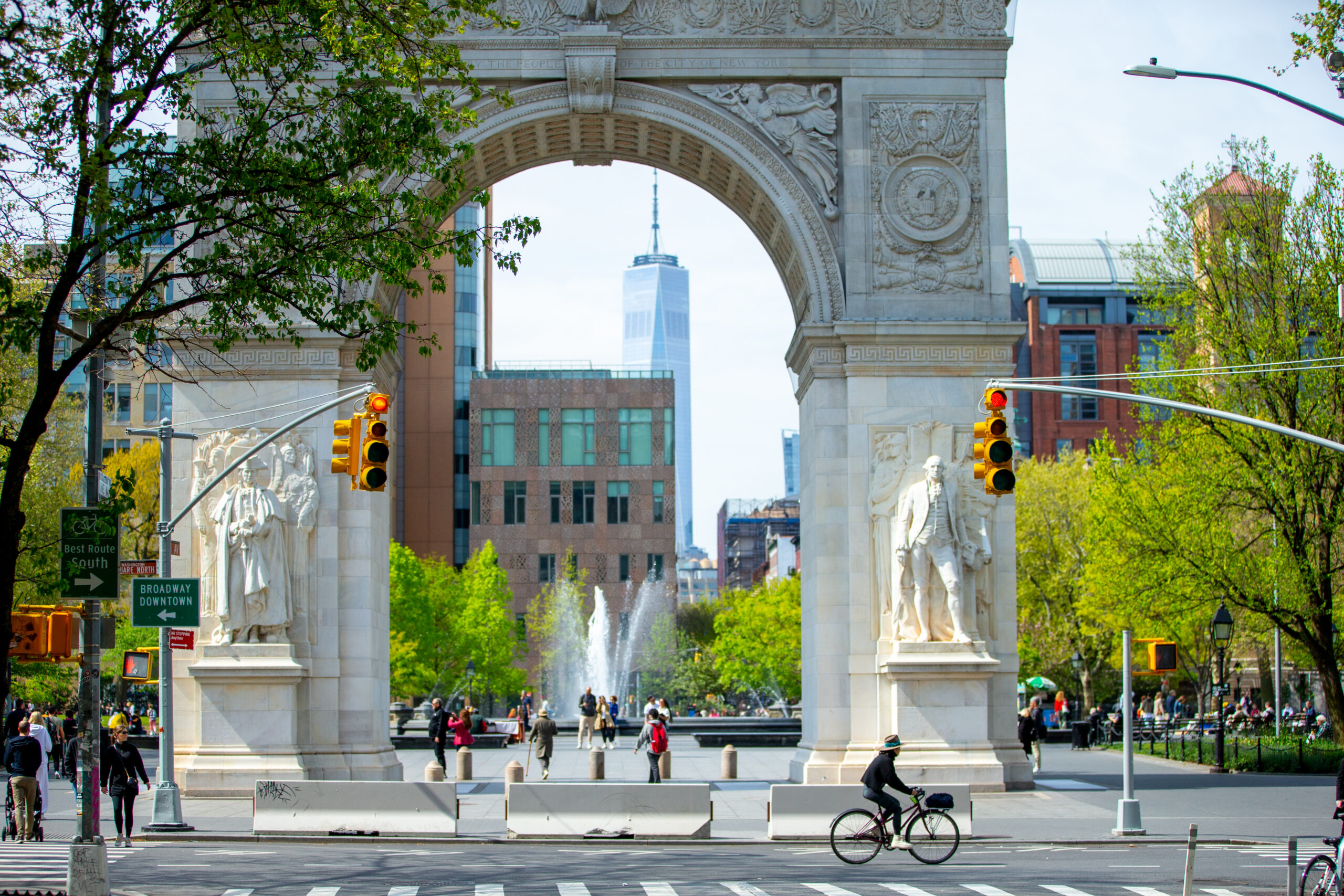 A person rides a bike past the Washington Square Arch in New York City, with the fountain and One World Trade Center visible in the background.