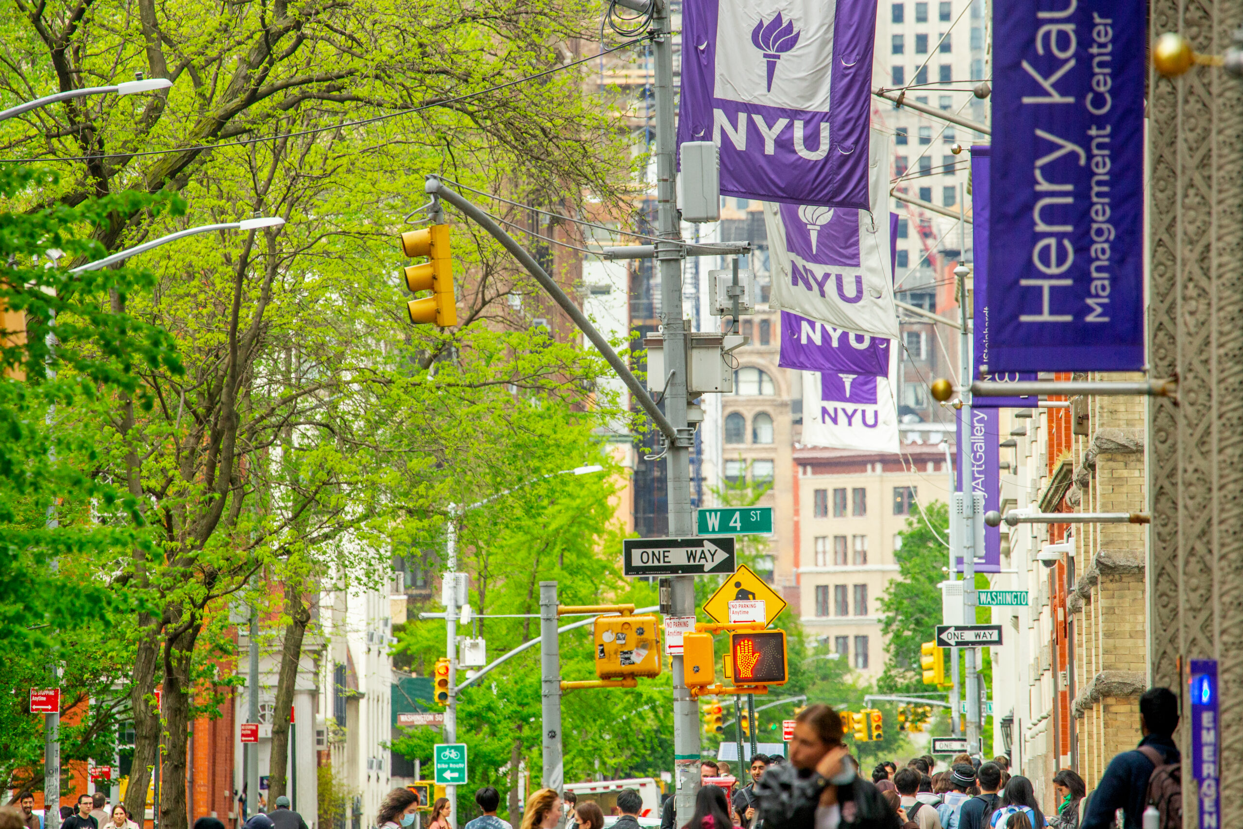 A busy street on the NYU campus, with large groups of people walking by, NYU flags waving above them.