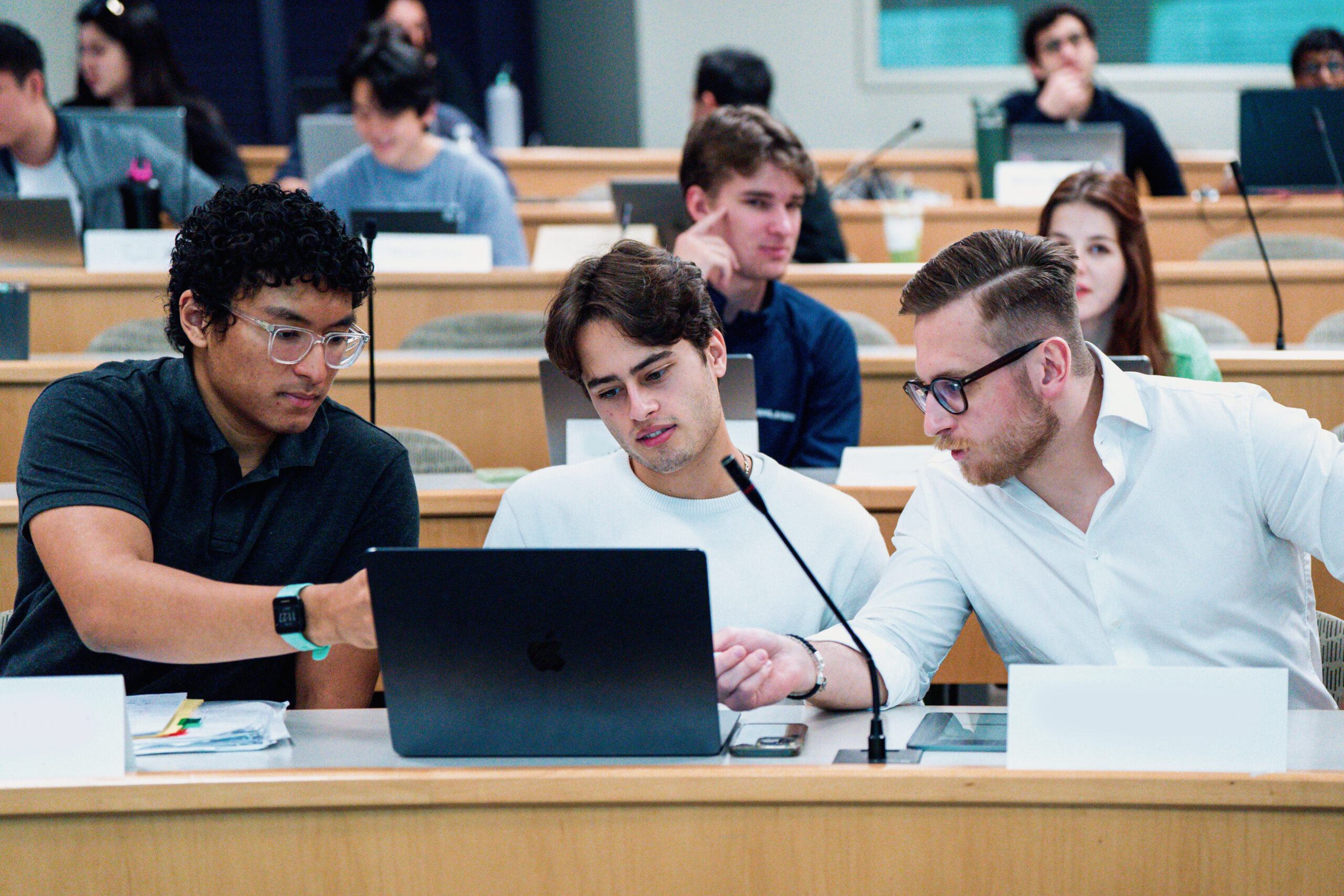 A group of students work together on a laptop in a lecture hall filled with other students.
