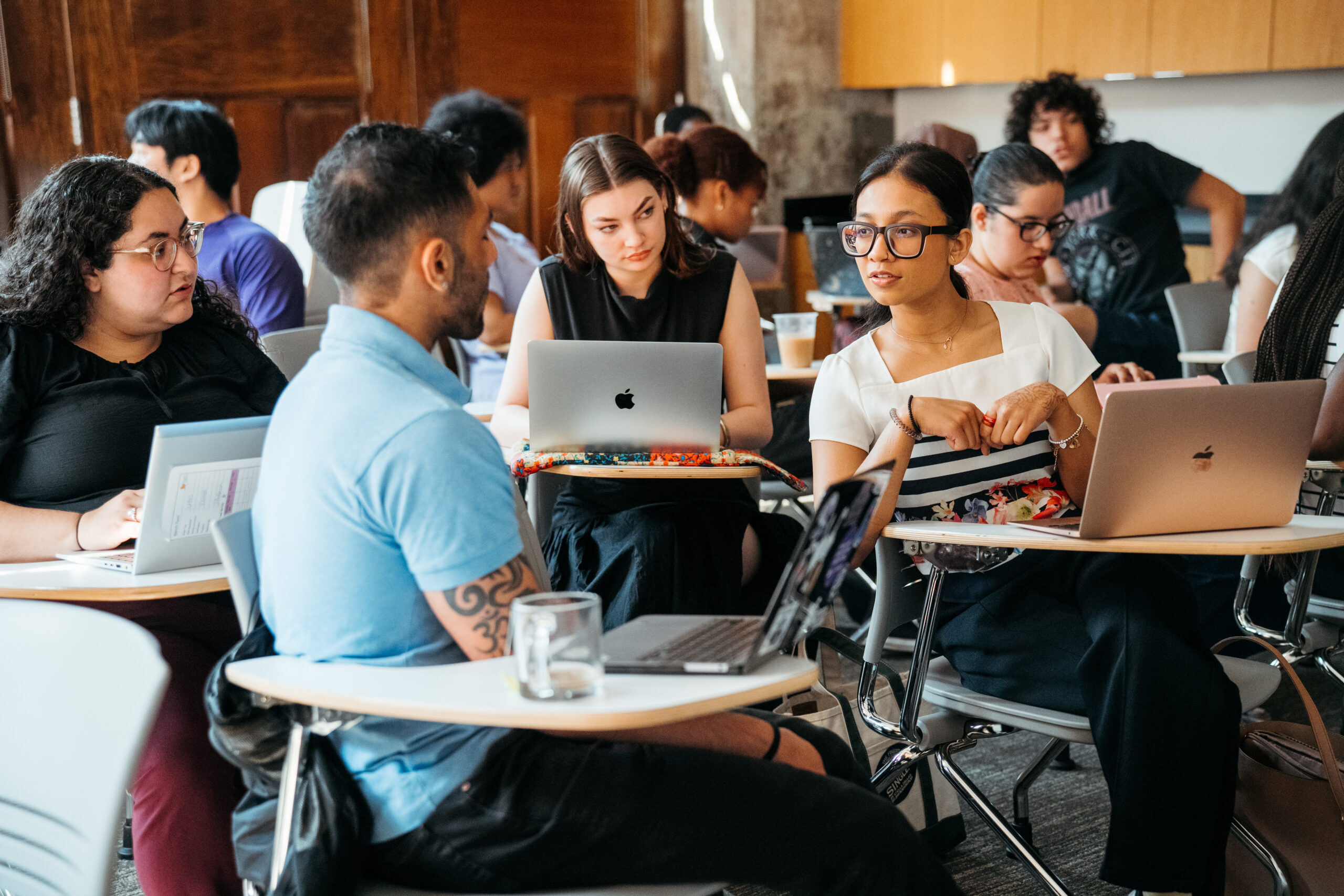 A group of students sit at desks with notebooks and laptops, chatting together in a small circle.