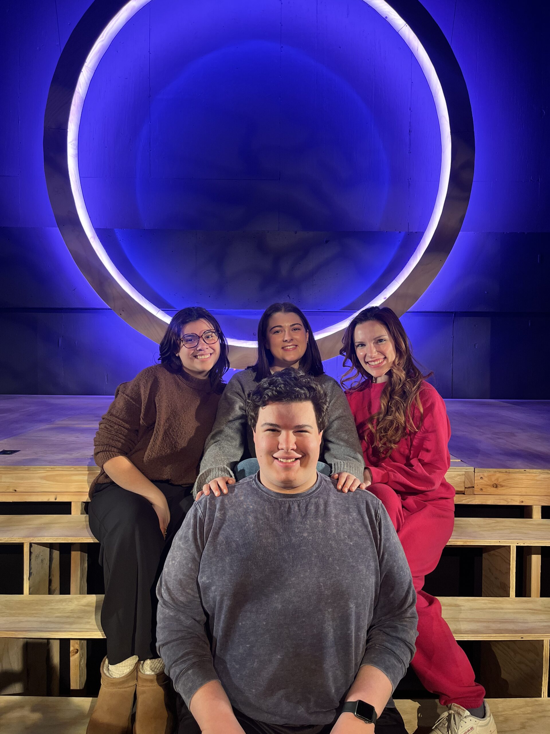 A group of Educational Theatre students sitting on a stage infront of a glowing ring.