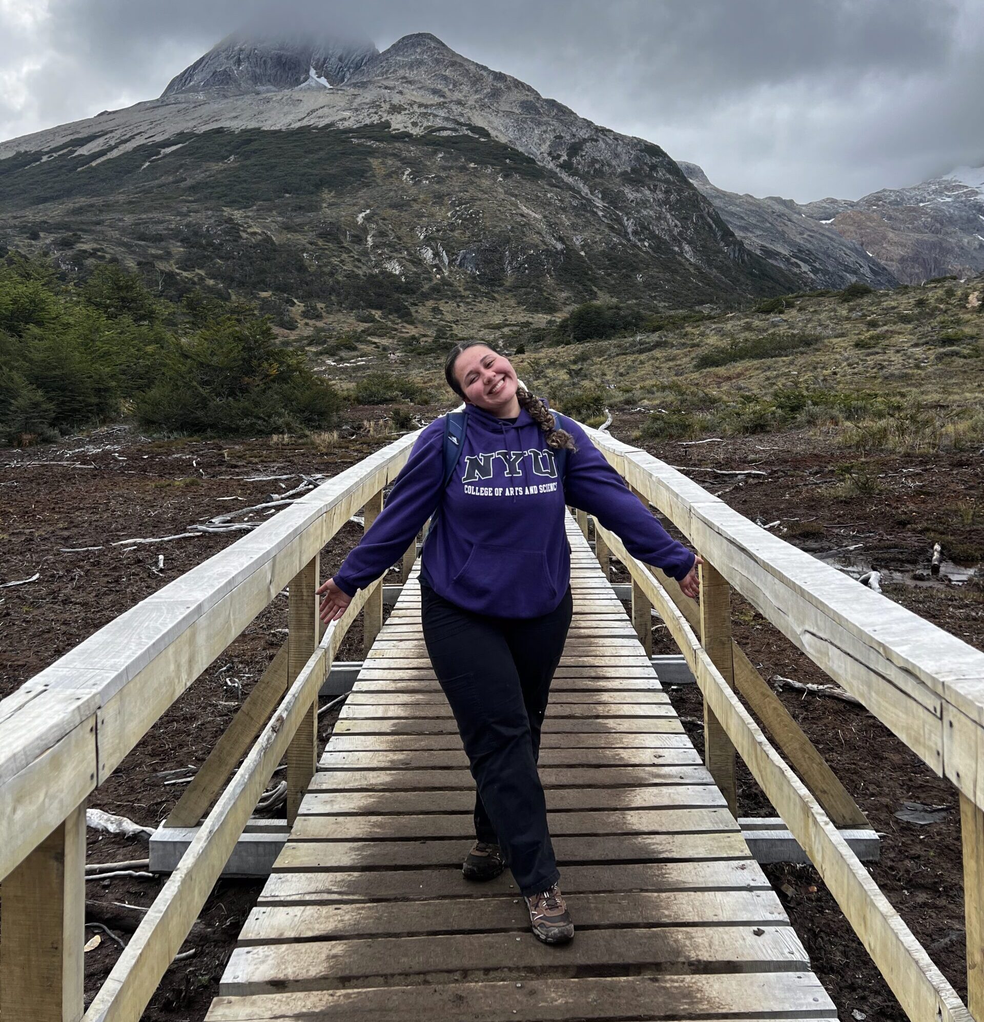 Yulianna Acuña stands in an NYU sweatshirt on a wooden bridge with a rocky peak rising into the clouds behind her.