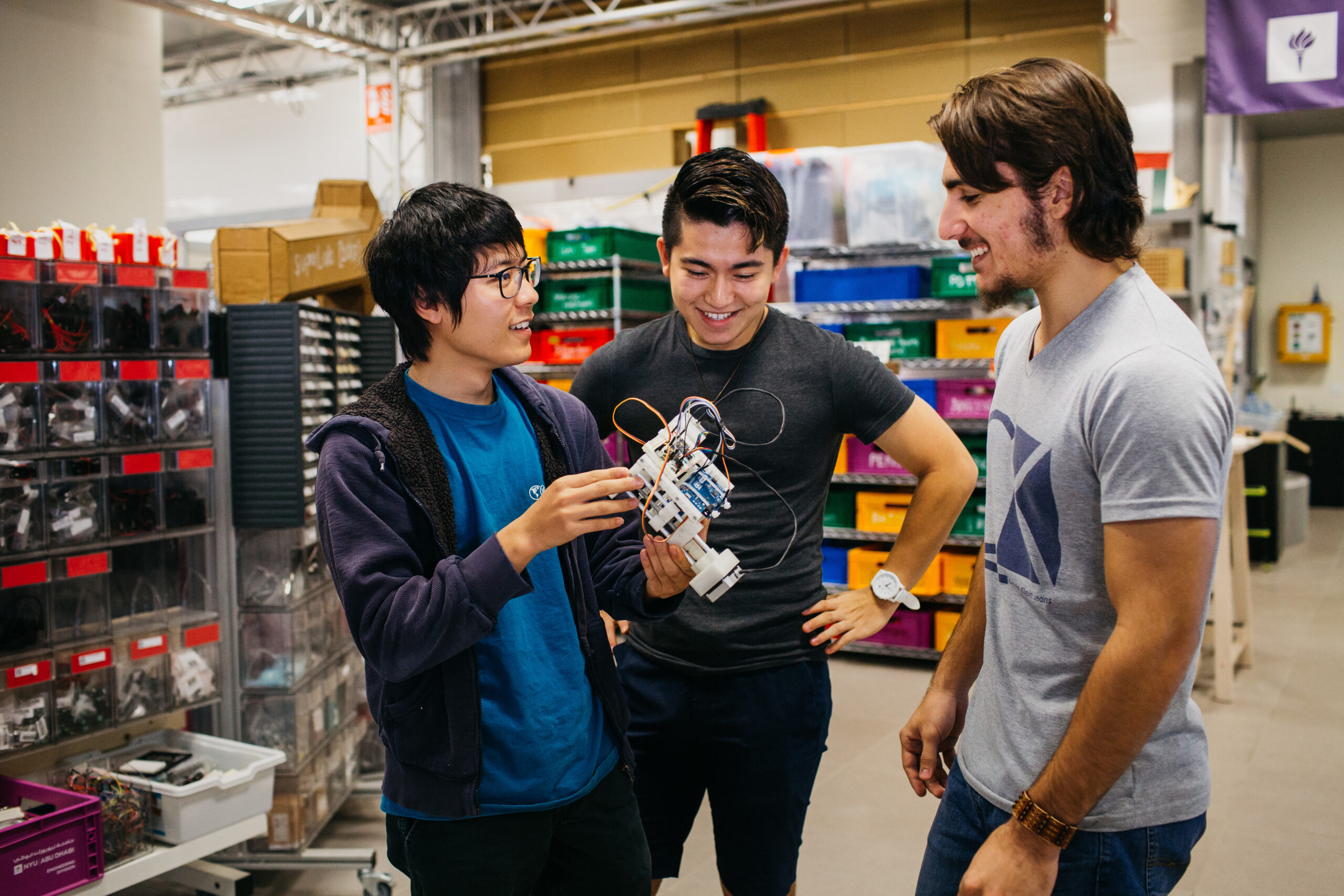 Three students in a lab smile and examine a small robot prototype together, surrounded by colorful bins and electronic components.