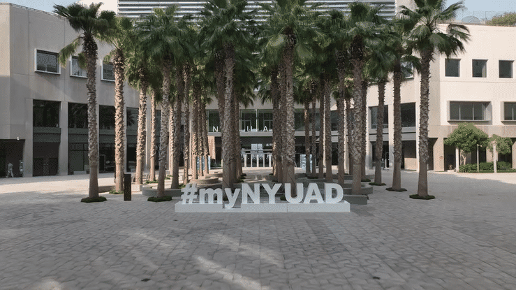 A drone shot moves through a row of palm trees toward a plaza with large white letters spelling “#myNYUAD” in front of a modern campus building.