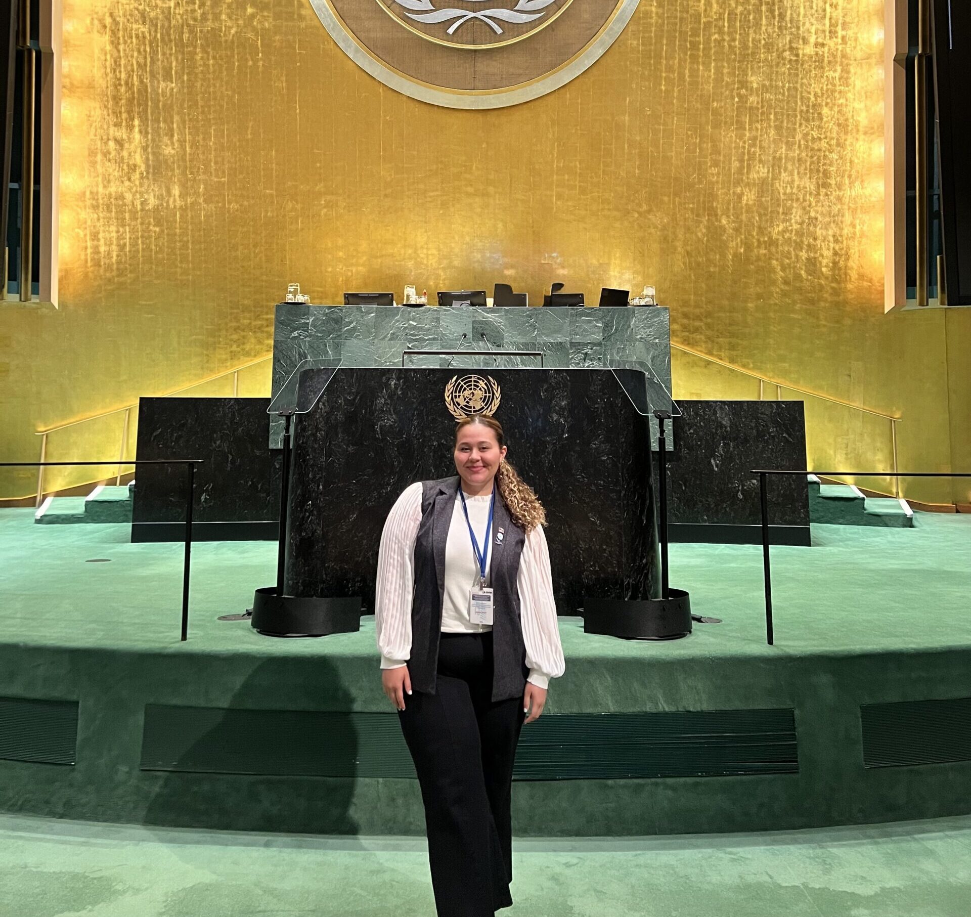 Yulianna Acuña stands, smiling, in front of the United Nations General Assembly podium, the UN emblem visible behind her.