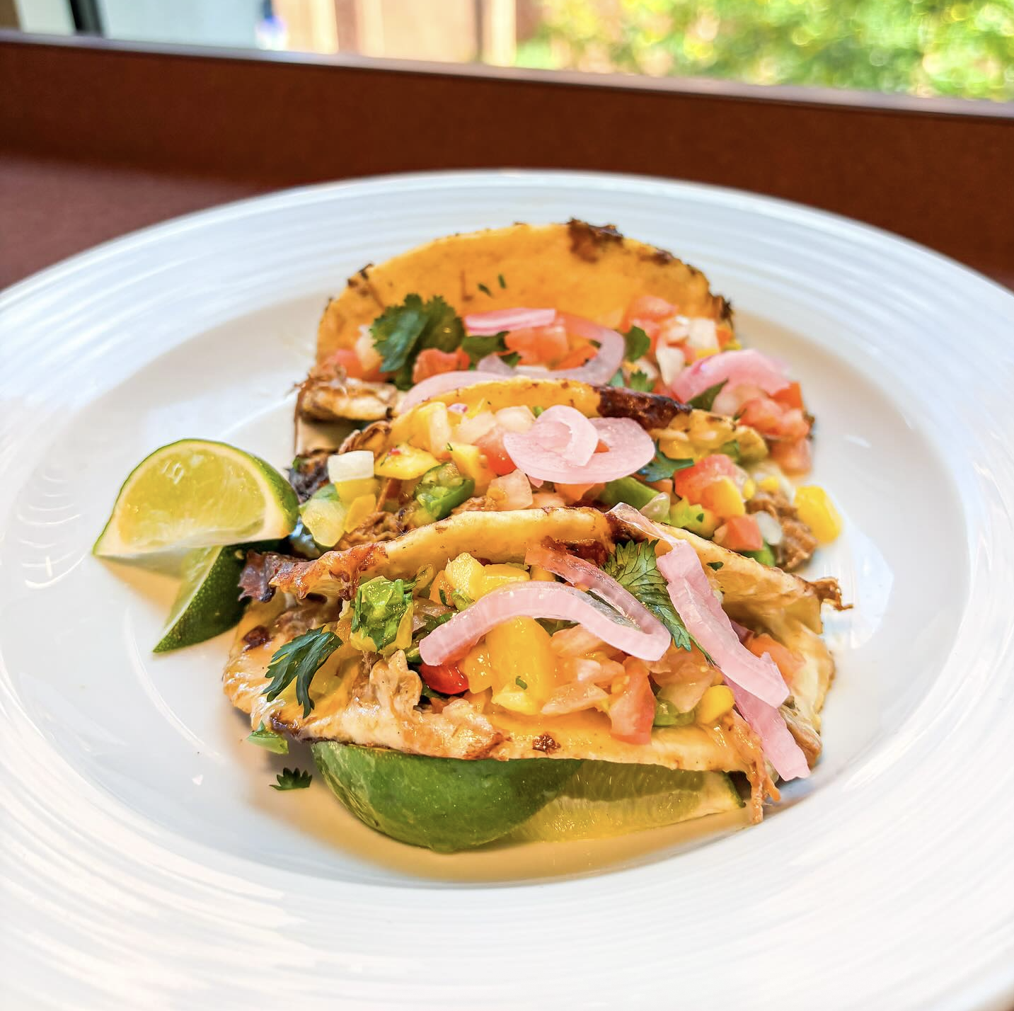 Three tacos filled with shredded meat, mango salsa, avocado, cilantro, and pickled onions served on a white plate with lime wedges.