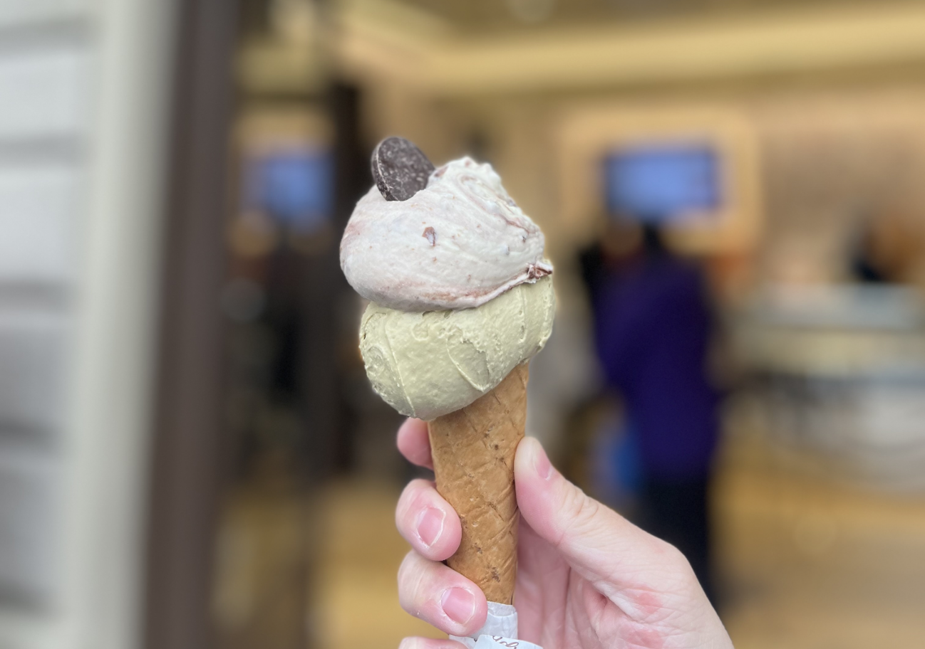 A close-up of a hand holding a cone of gelato with the storefront out of focus in the background.