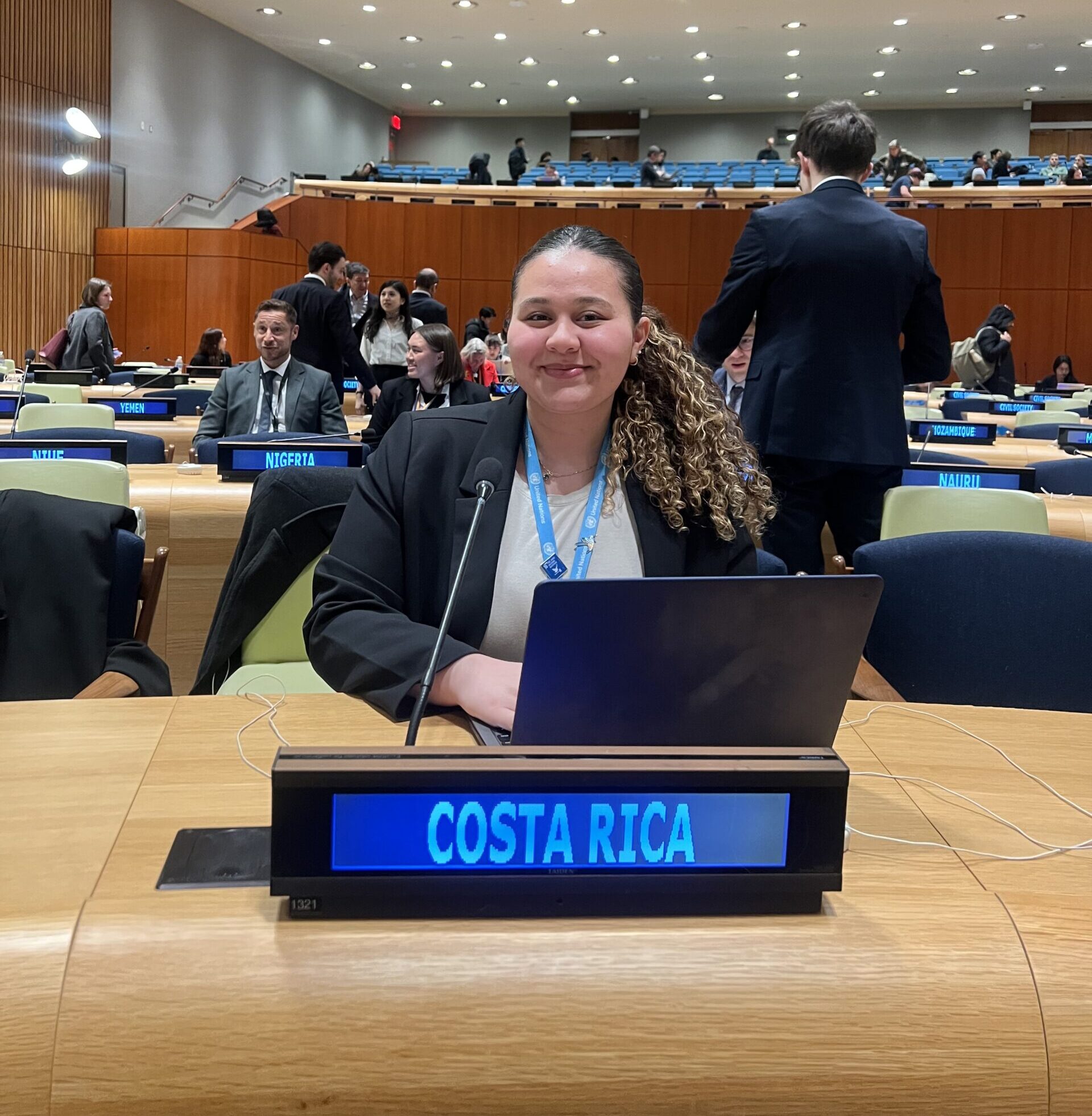 Smiling, Yulianna Acuña sits at a desk with a laptop in front of a sign that reads “Costa Rica” at the United Nations.