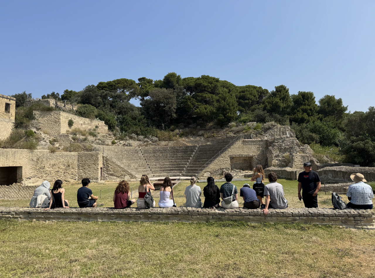 A group of students sit in a row facing the ruins of an ancient Roman theater surrounded by greenery under a clear sky.
