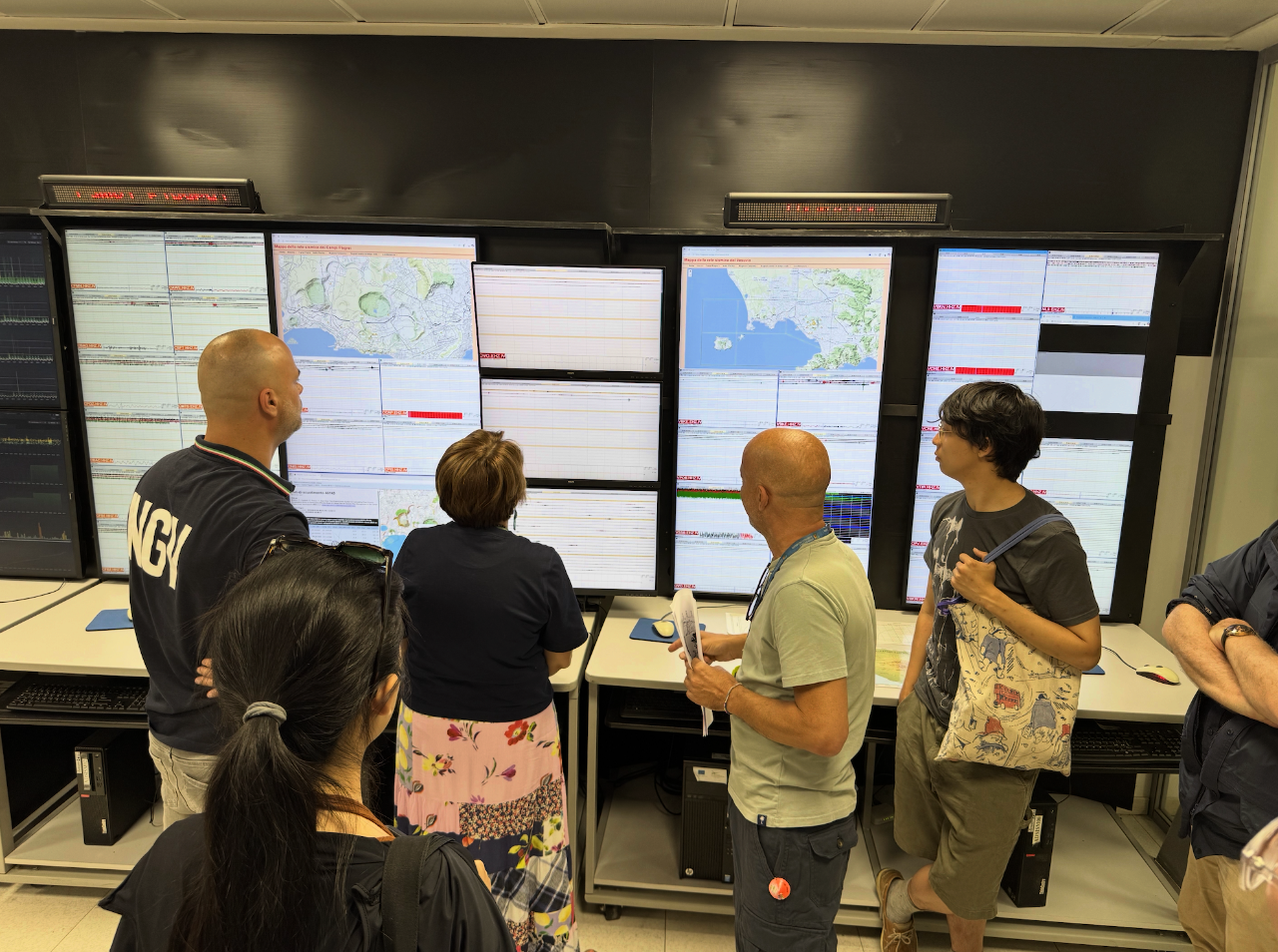 A group of people stand in front of large screens displaying maps and seismic data inside the monitoring room at the Vesuvius Observatory