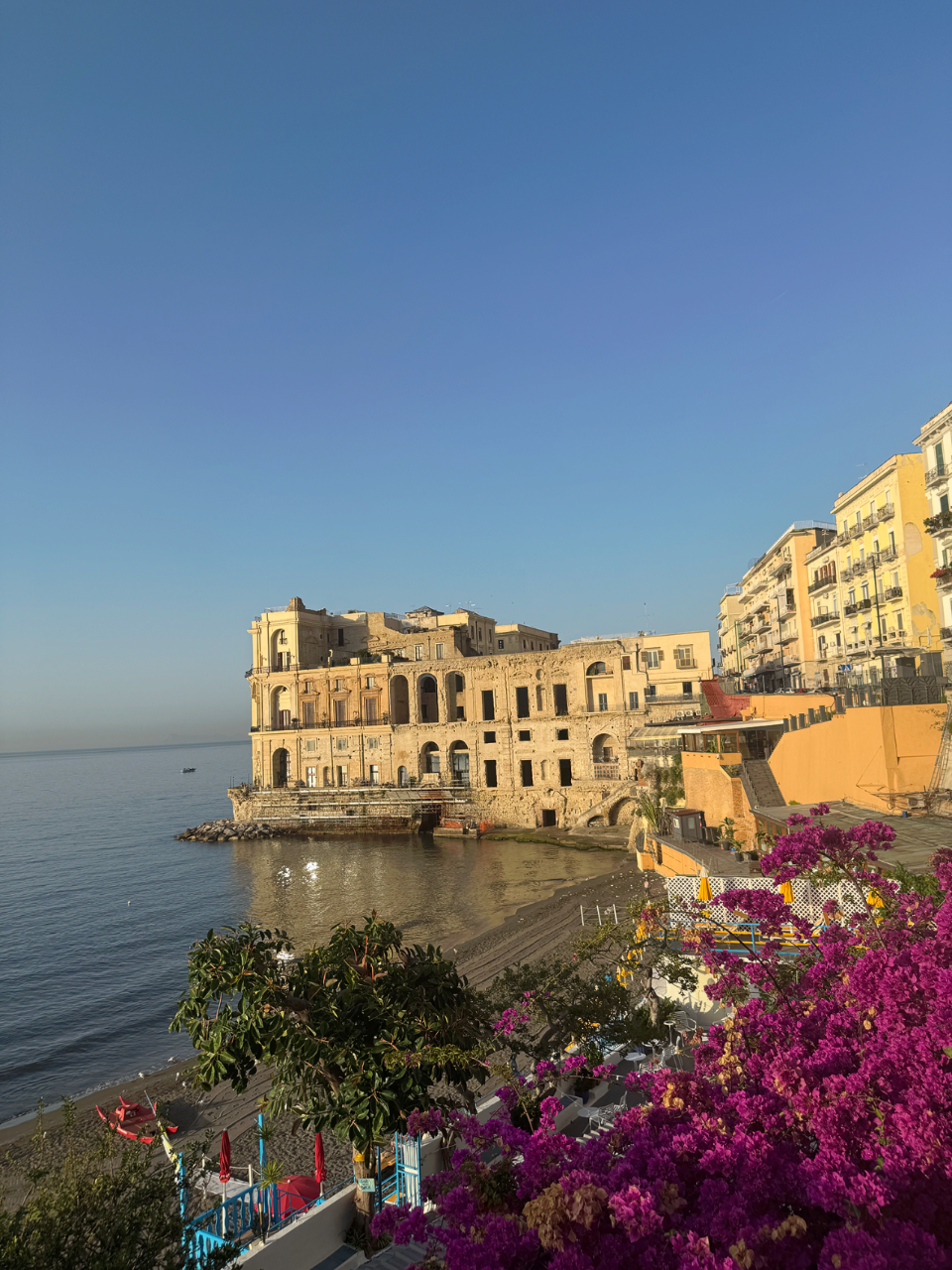 A coastal Italian town with an old stone building overlooking calm blue water, with colorful flowers and beach umbrellas in the foreground under a clear sky.